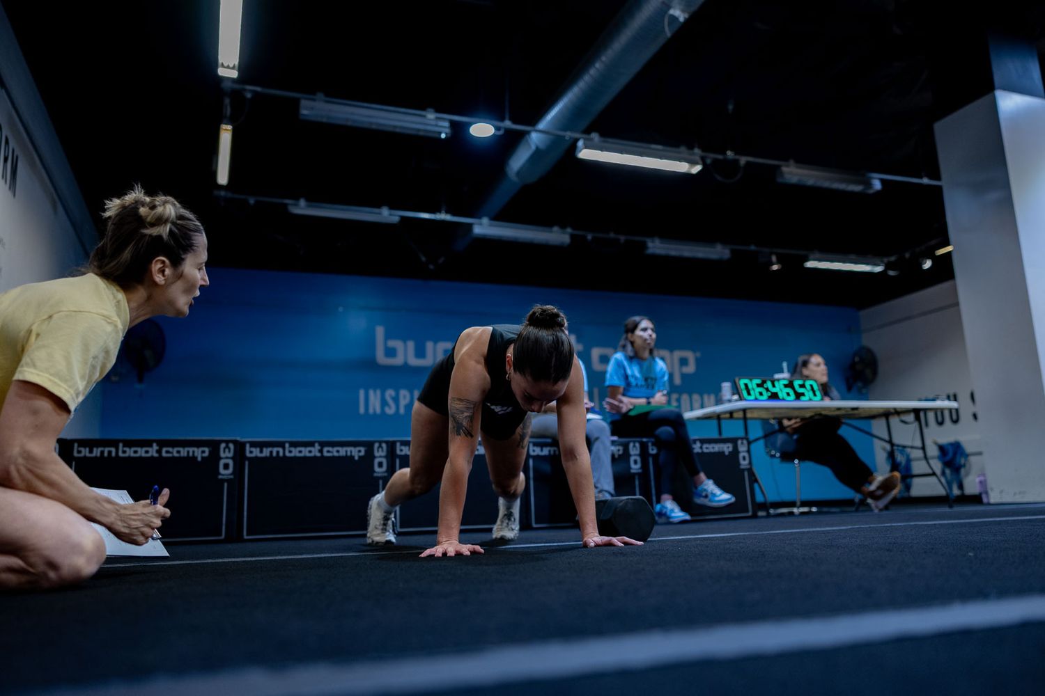 People in workout attire prepare at starting positions on a gym floor with coaches looking on during a fitness class.