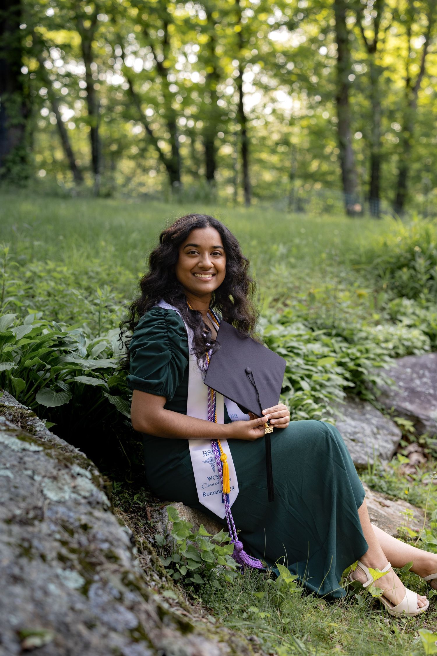 Graduate in dark green dress sits on rock in forest setting holding graduation cap with honors cords.