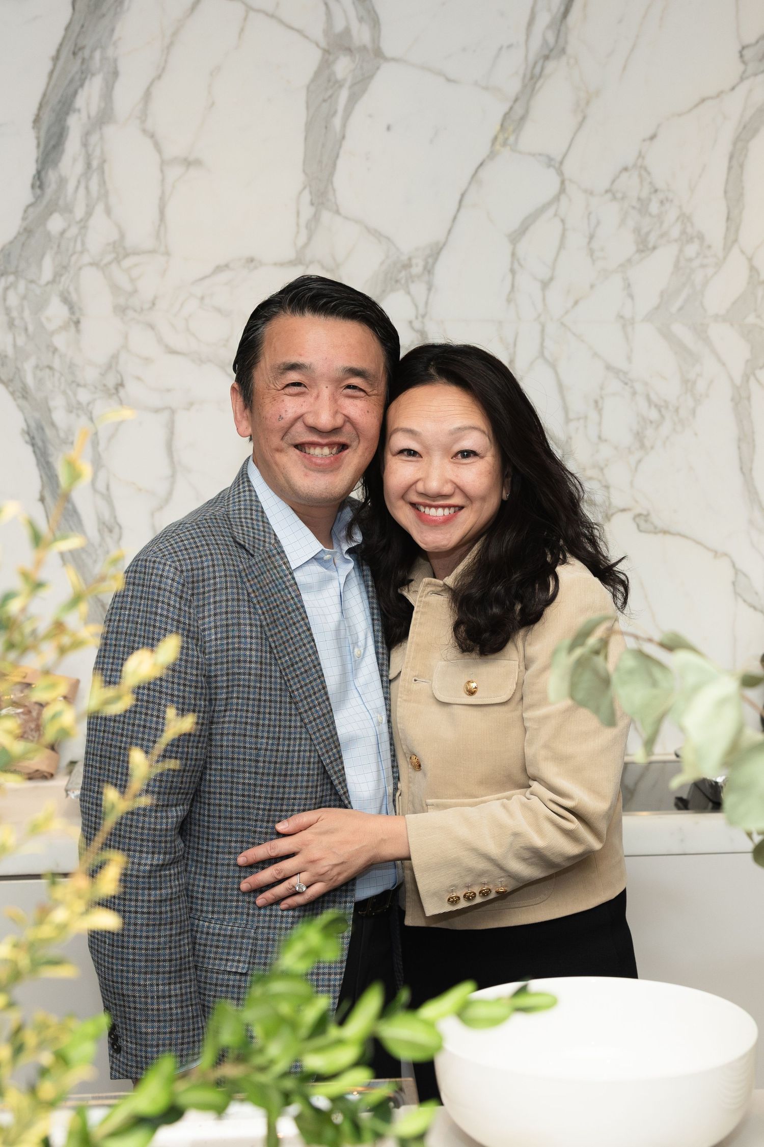 Two people embrace and smile warmly in an elegant setting with marble walls and decorative plants.