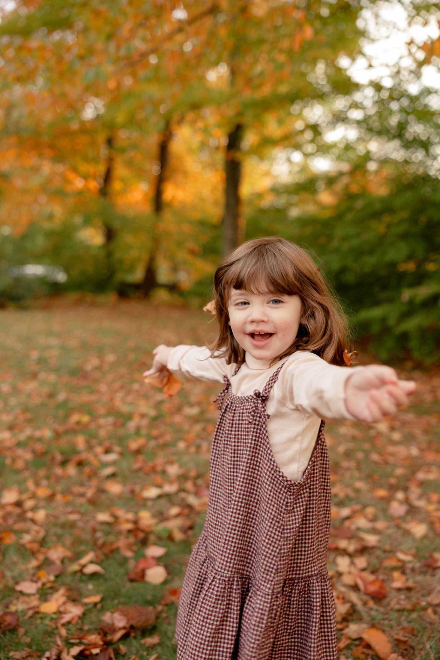 Child in brown gingham dress plays joyfully among fallen autumn leaves in a park setting.
