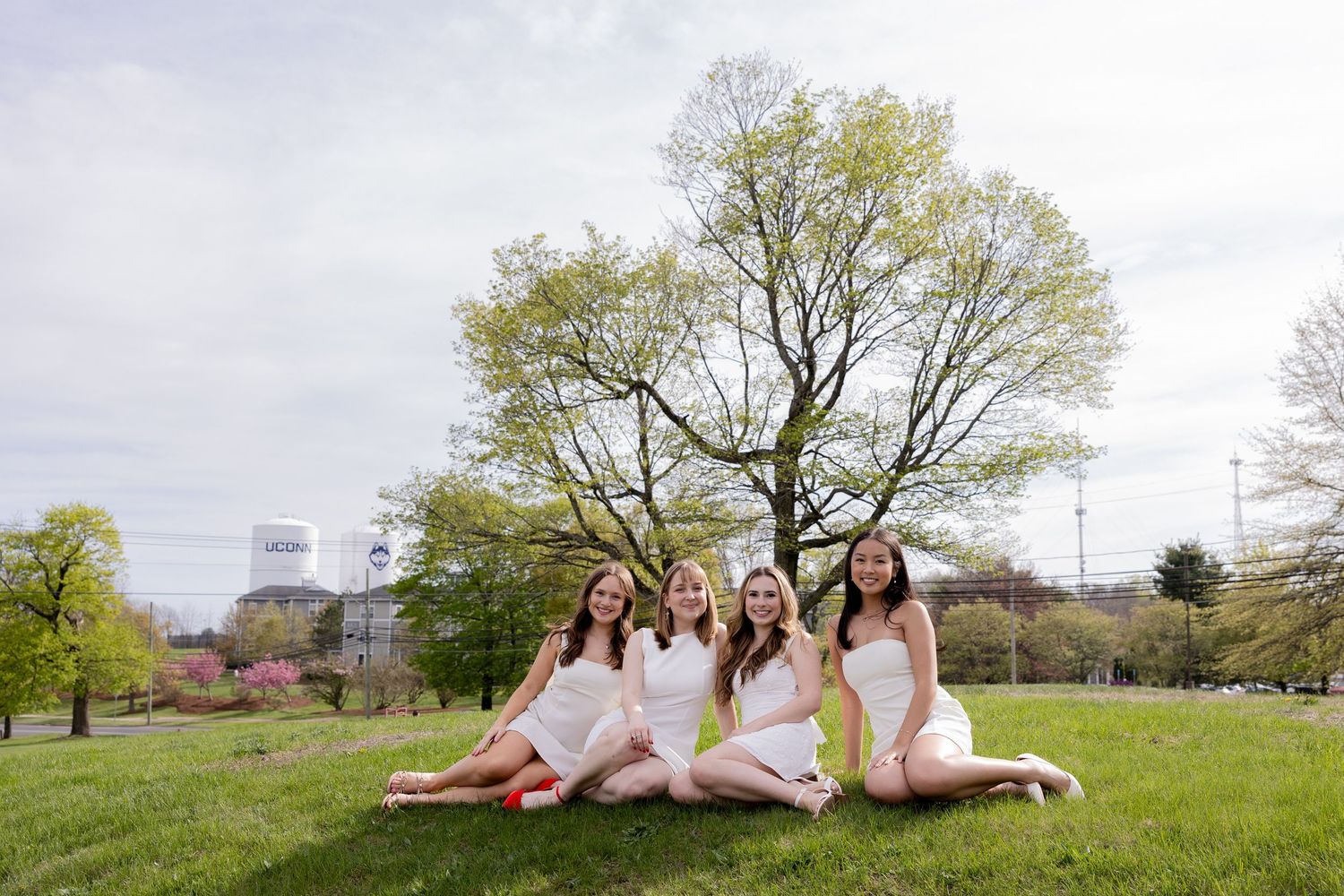 Four women in white dresses sit together on green grass with spring trees and buildings in the background.