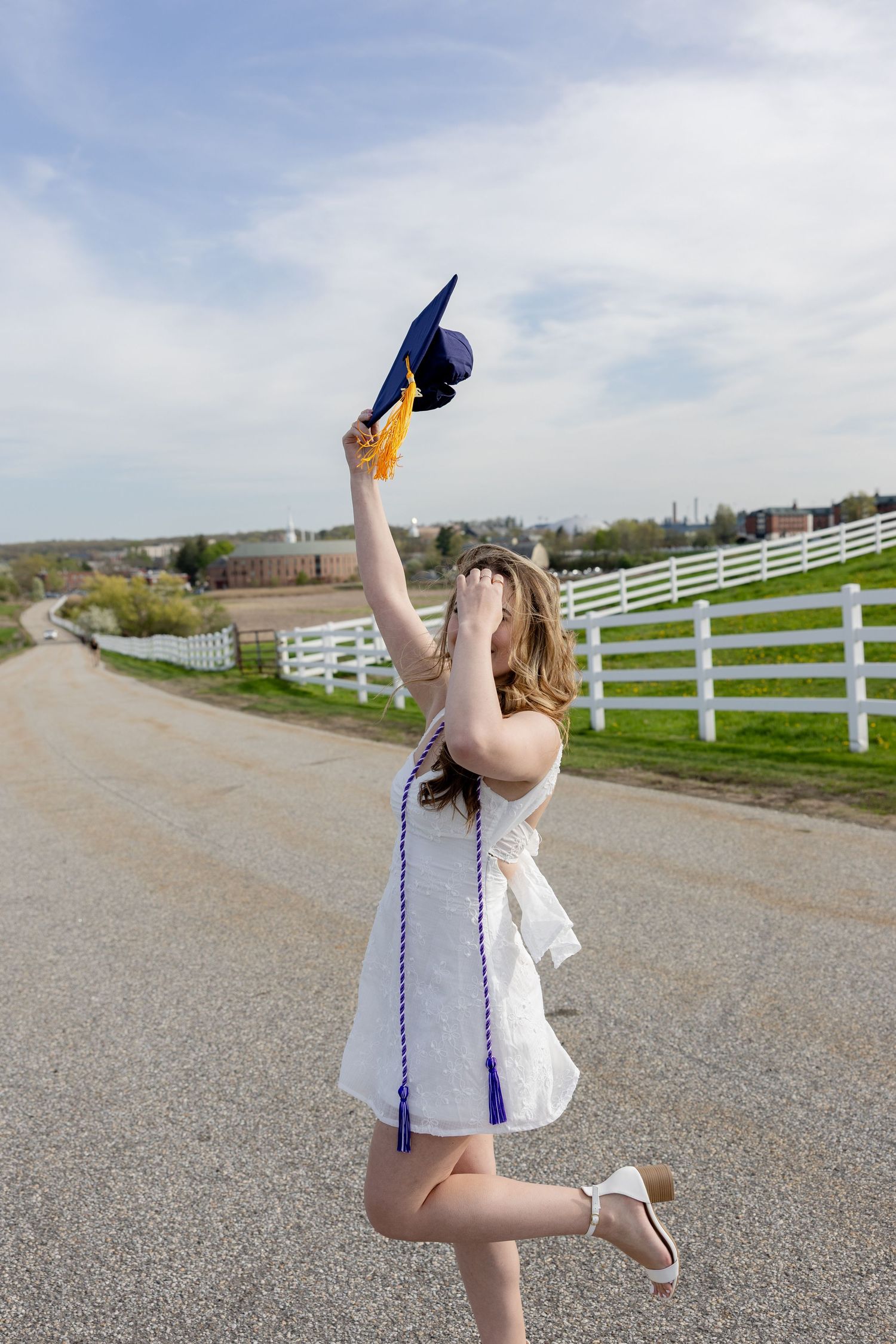 Graduate in white dress celebrates outdoors along dirt road with white fence, holding blue graduation cap up.