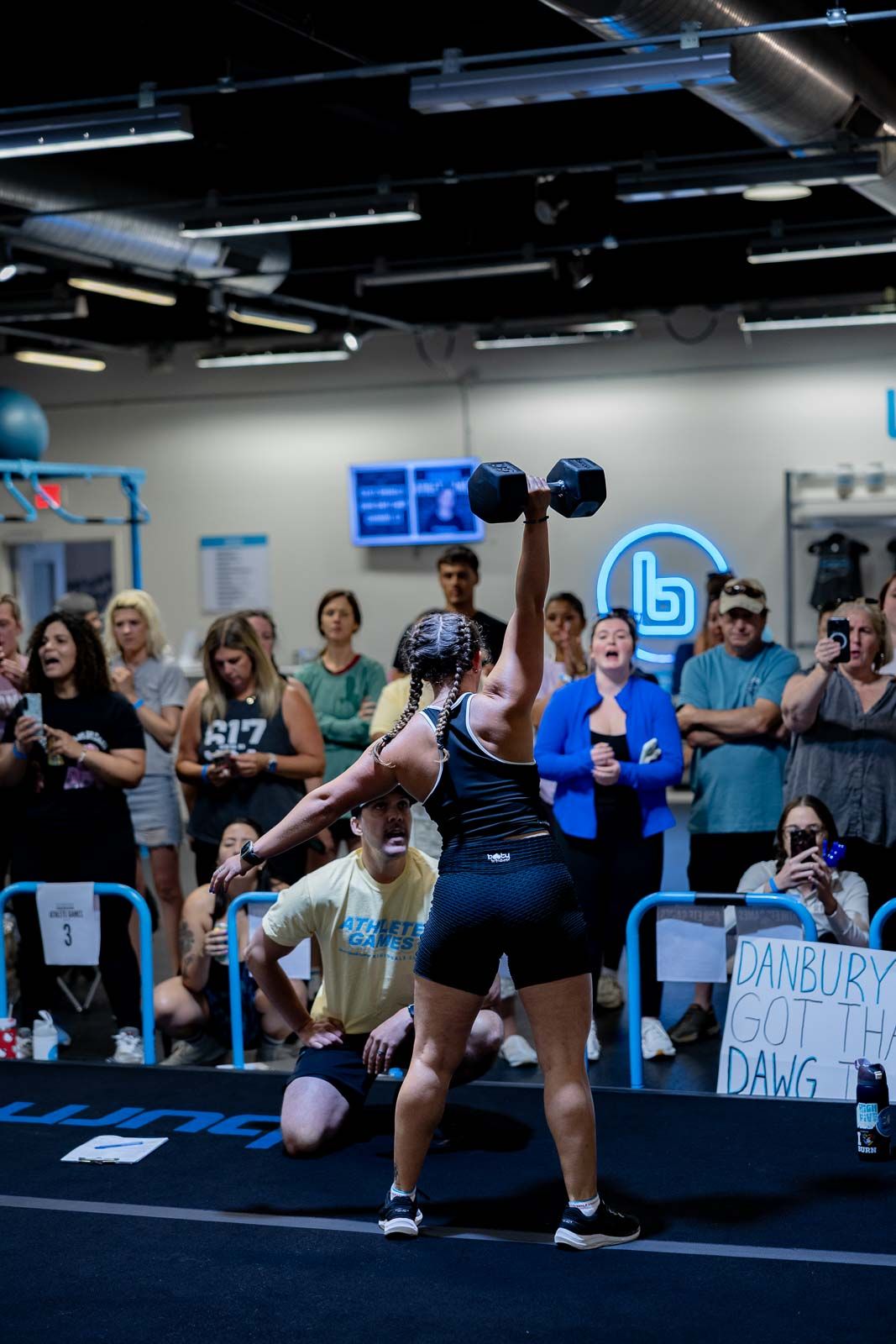 Athletes line up along a gym wall during a CrossFit competition while being watched by spectators in the background.