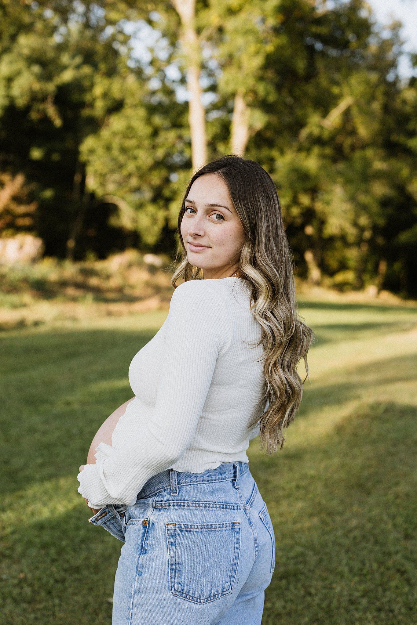Pregnant woman in white top and jeans poses outdoors with long wavy hair.