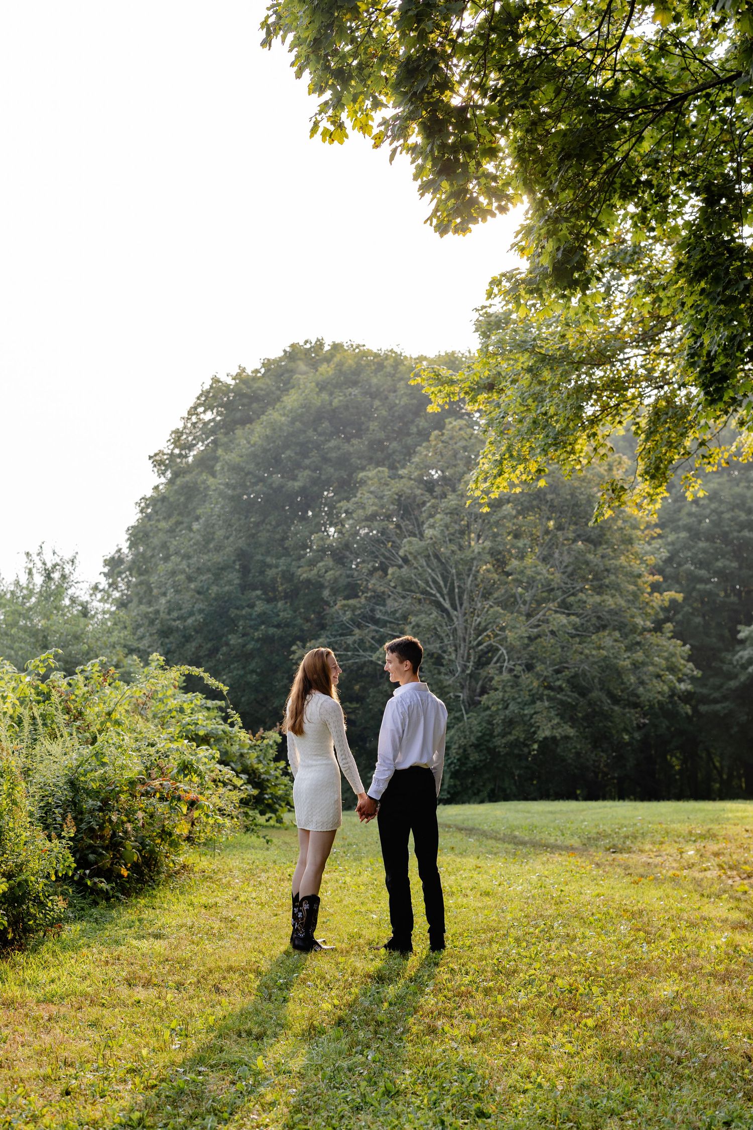 A couple stands together in a sunlit meadow surrounded by lush green trees on a warm summer afternoon.