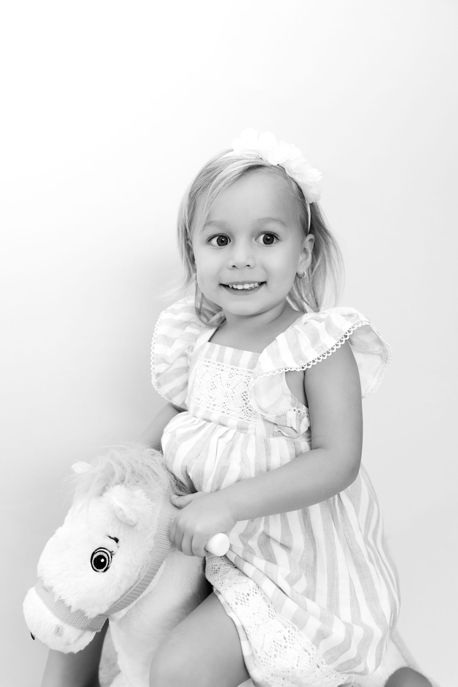 Black and white portrait of a young child in a white dress sitting with a plush toy horse.