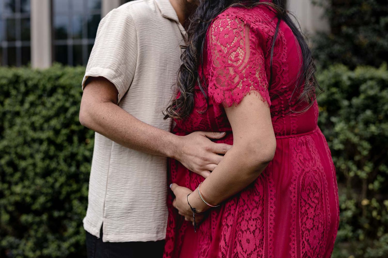 A couple embraces while standing in front of green hedges, with one person wearing a vibrant red lace dress.