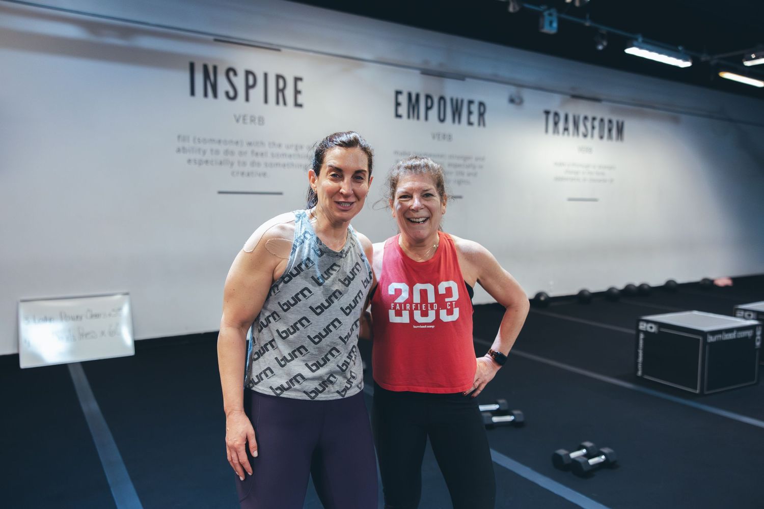 Two fitness enthusiasts posing together in front of motivational wall text at a gym.