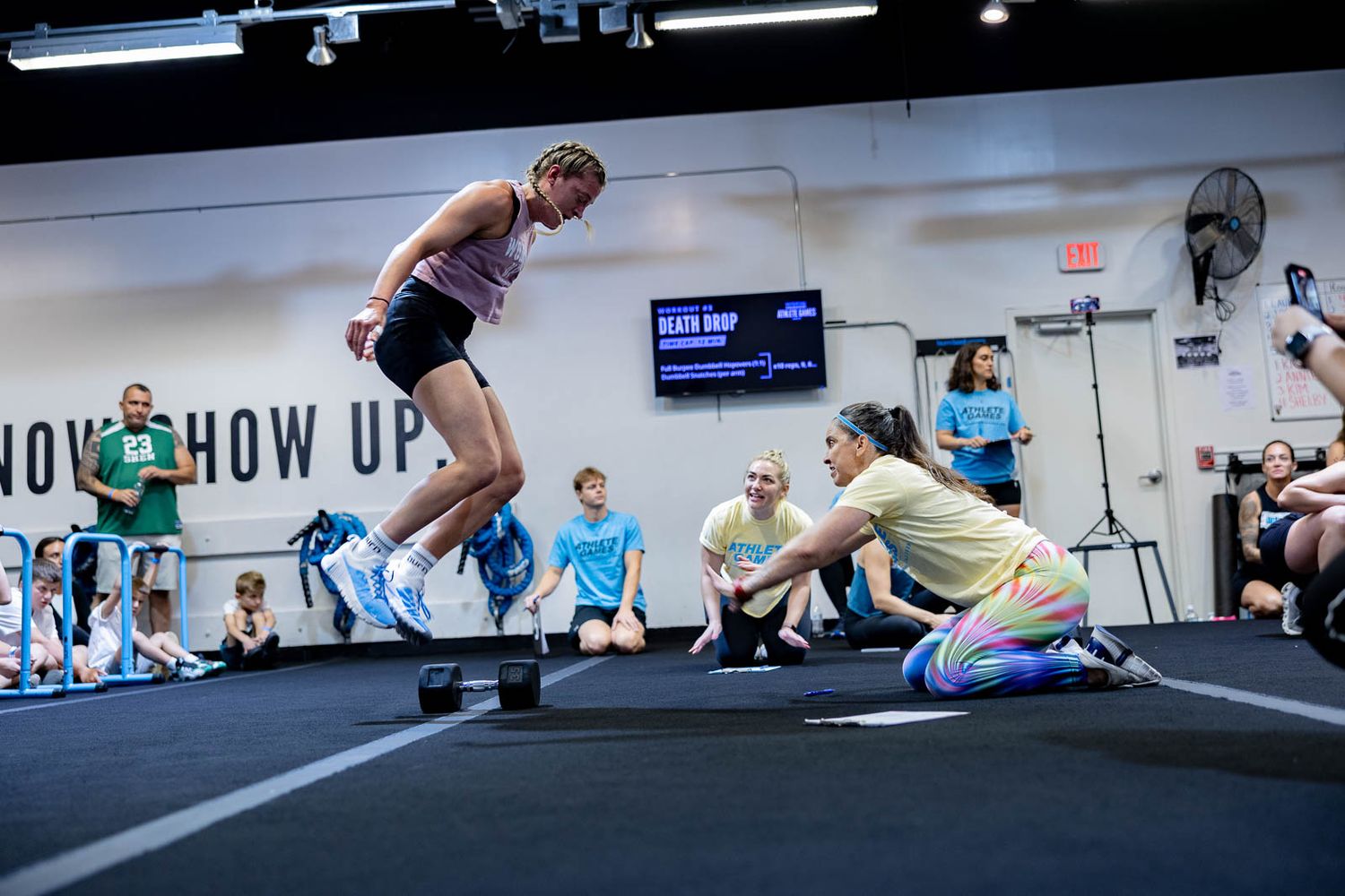 Group performs jumping burpees in sequence across a gym floor with fitness equipment visible.
