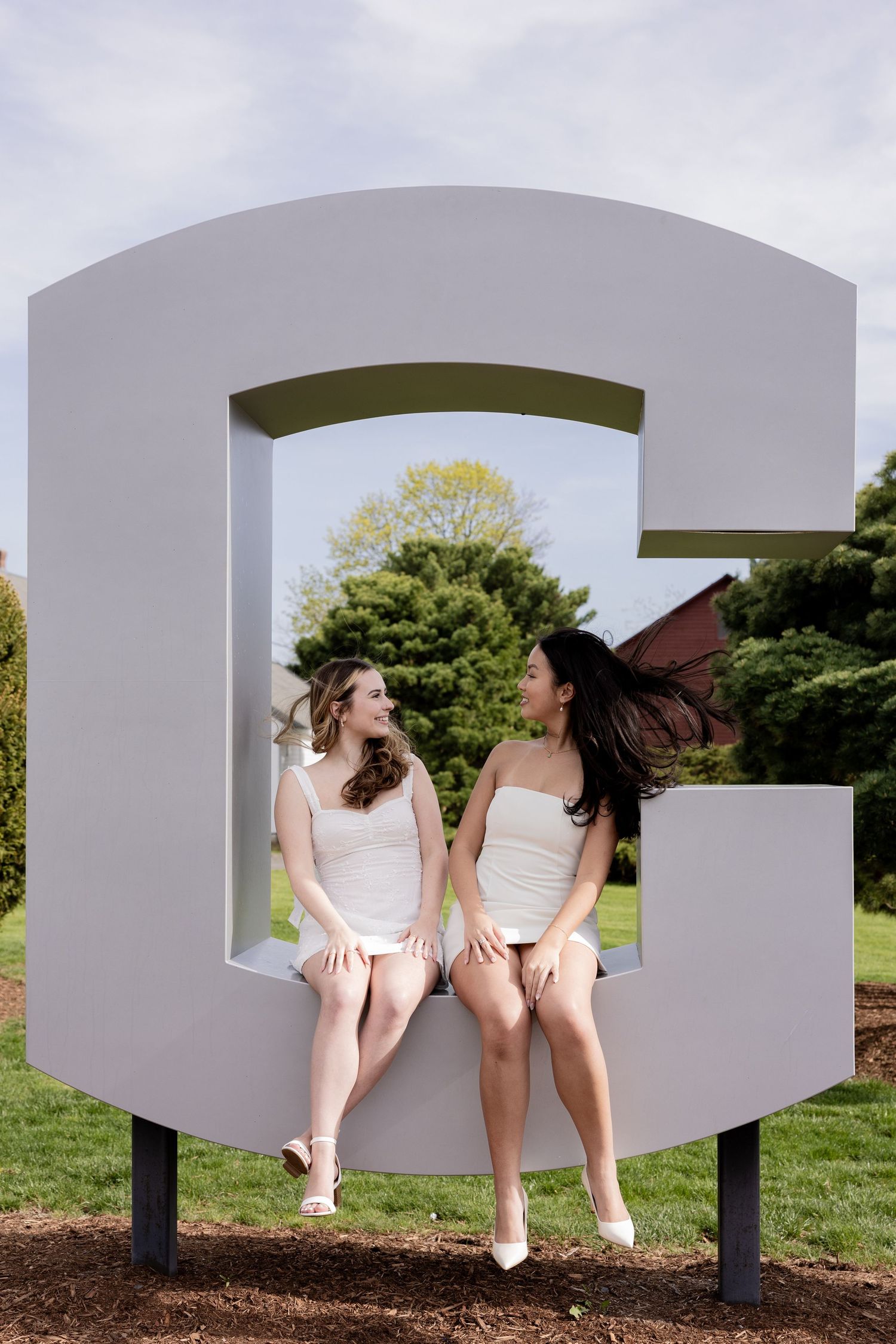 Two friends in white dresses sit together and laugh in a large decorative letter G sculpture outdoors.