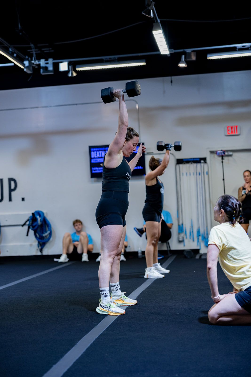 Multiple people performing overhead dumbbell exercises in a gym with 'UP' text visible on the wall.