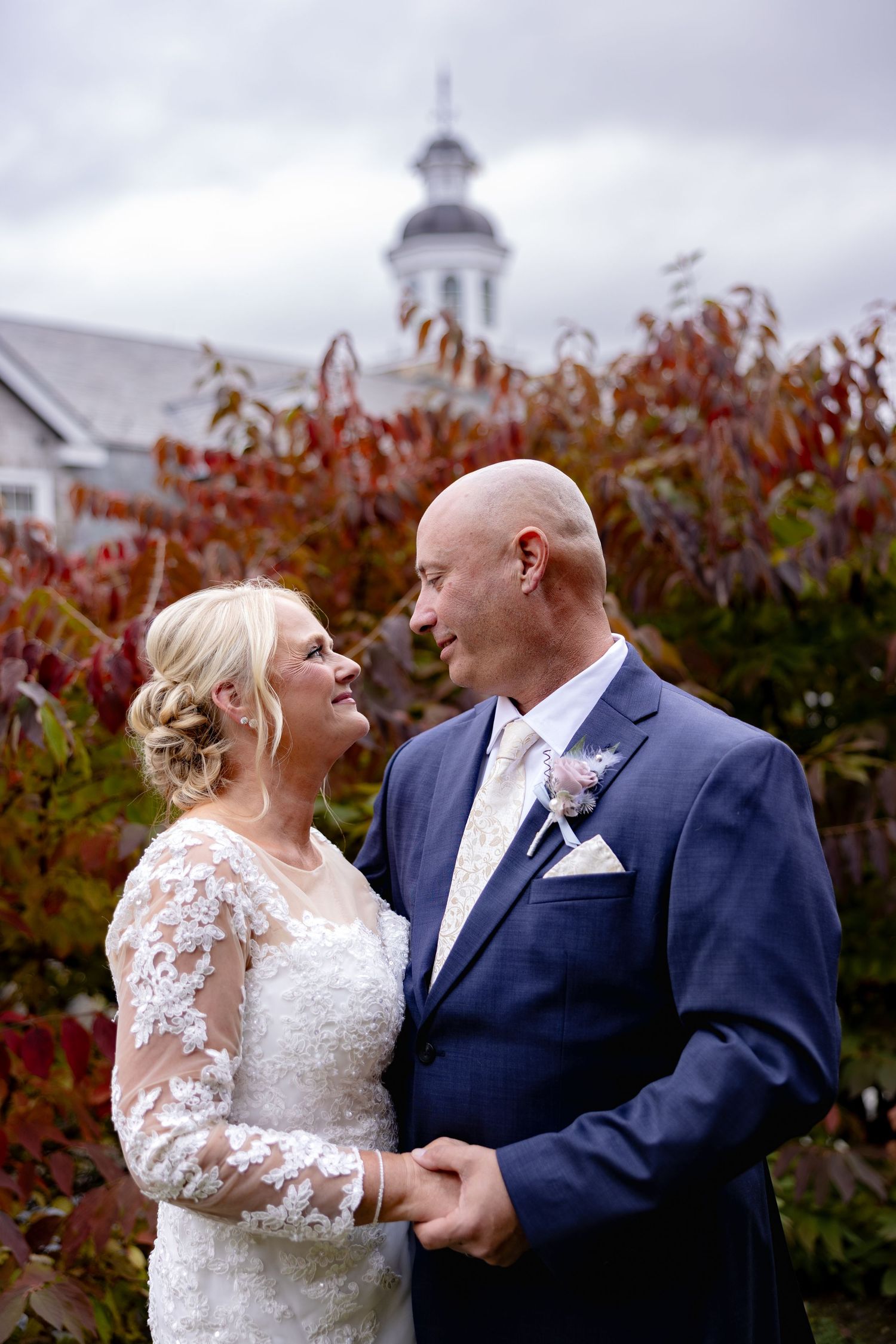A couple shares a tender moment in their wedding attire against an autumn backdrop with red foliage.