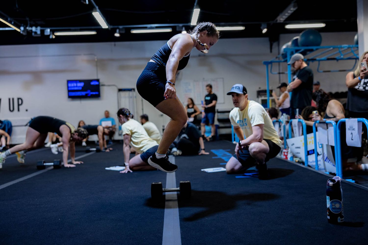 Group fitness class participants exercising together in a modern gym facility with blue accents and fluorescent lighting.