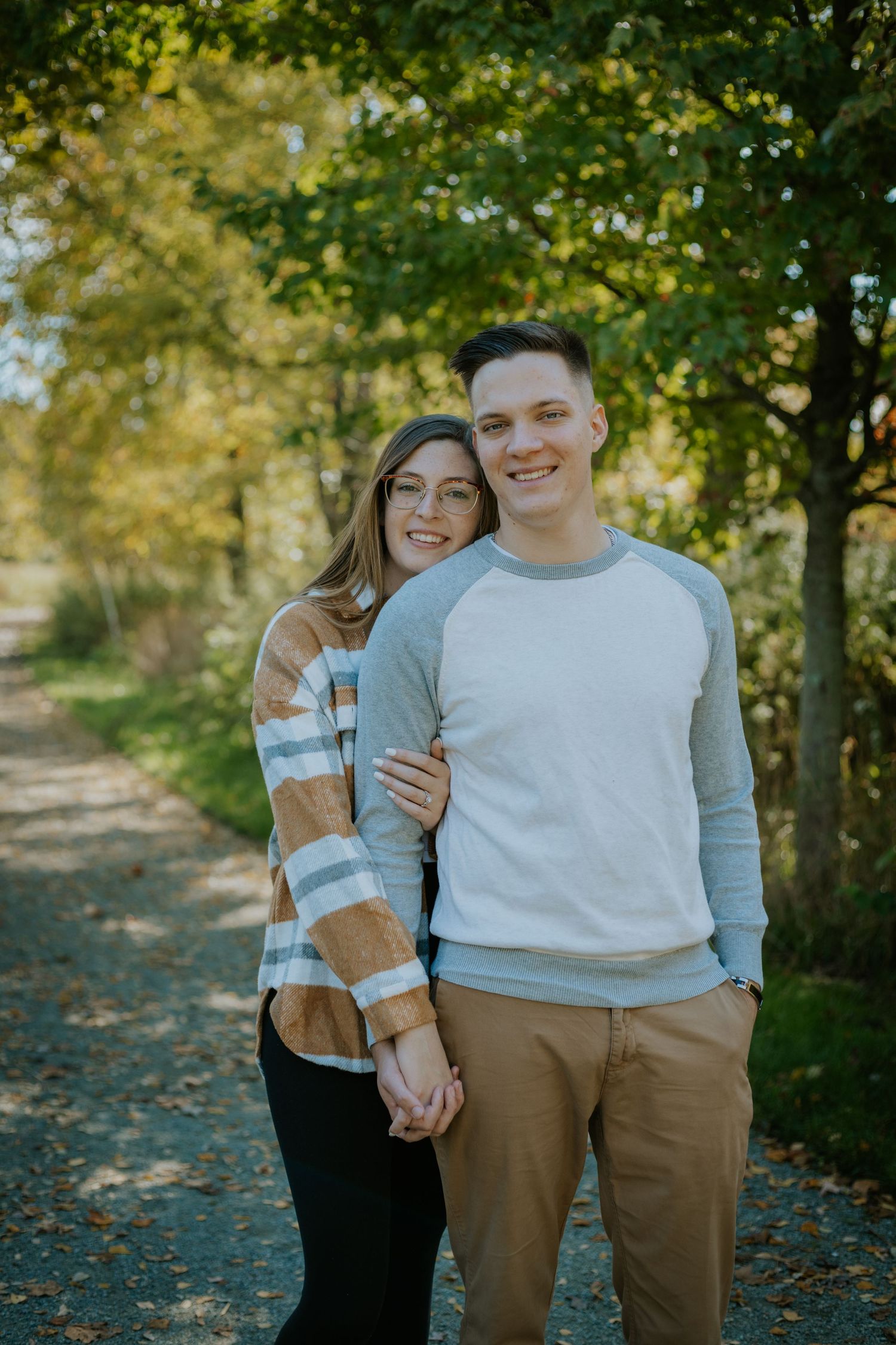 A couple poses together on a tree-lined autumn path wearing casual sweaters.