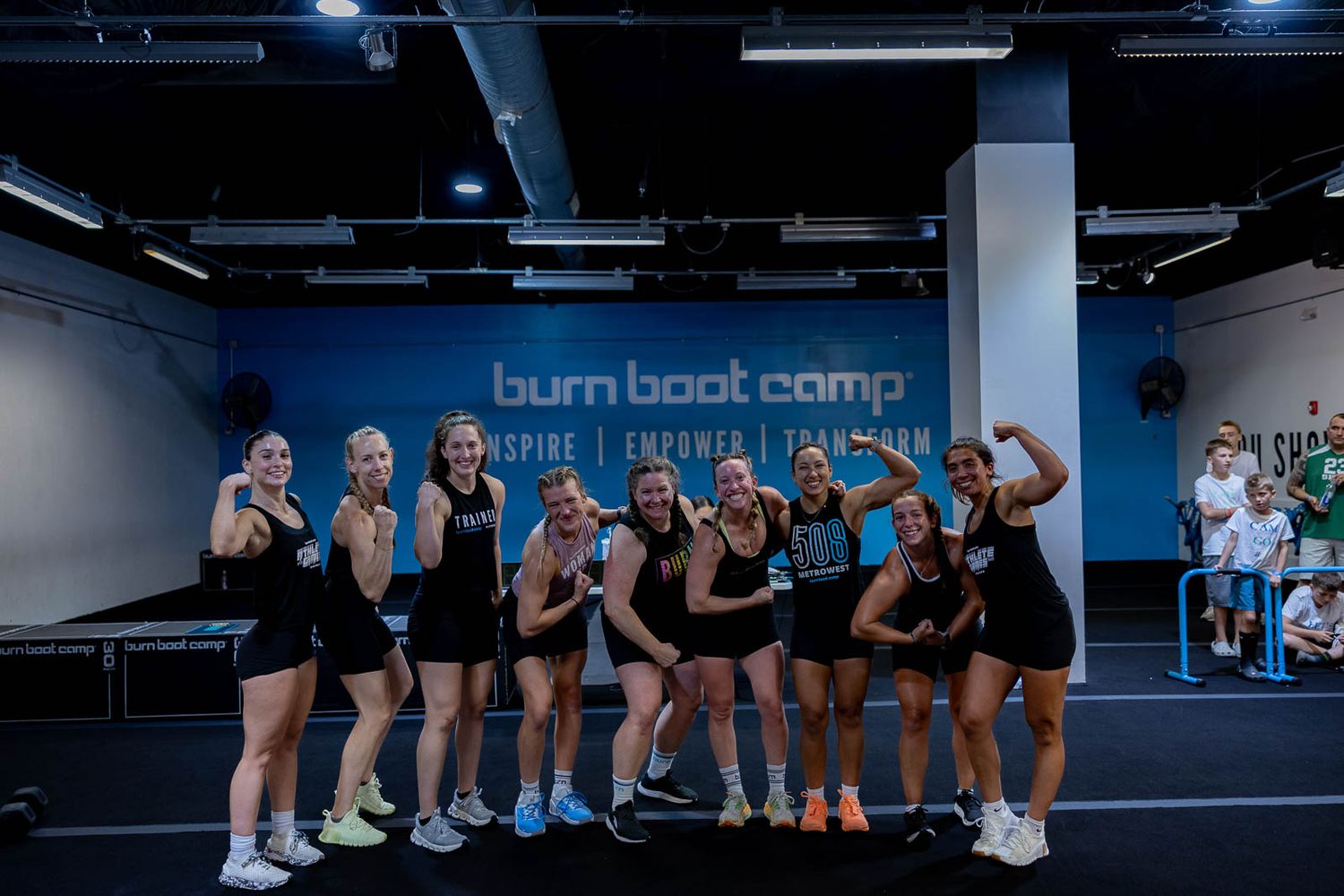 Group workout participants lined up in a dark gym space with blue floors and industrial ceiling.