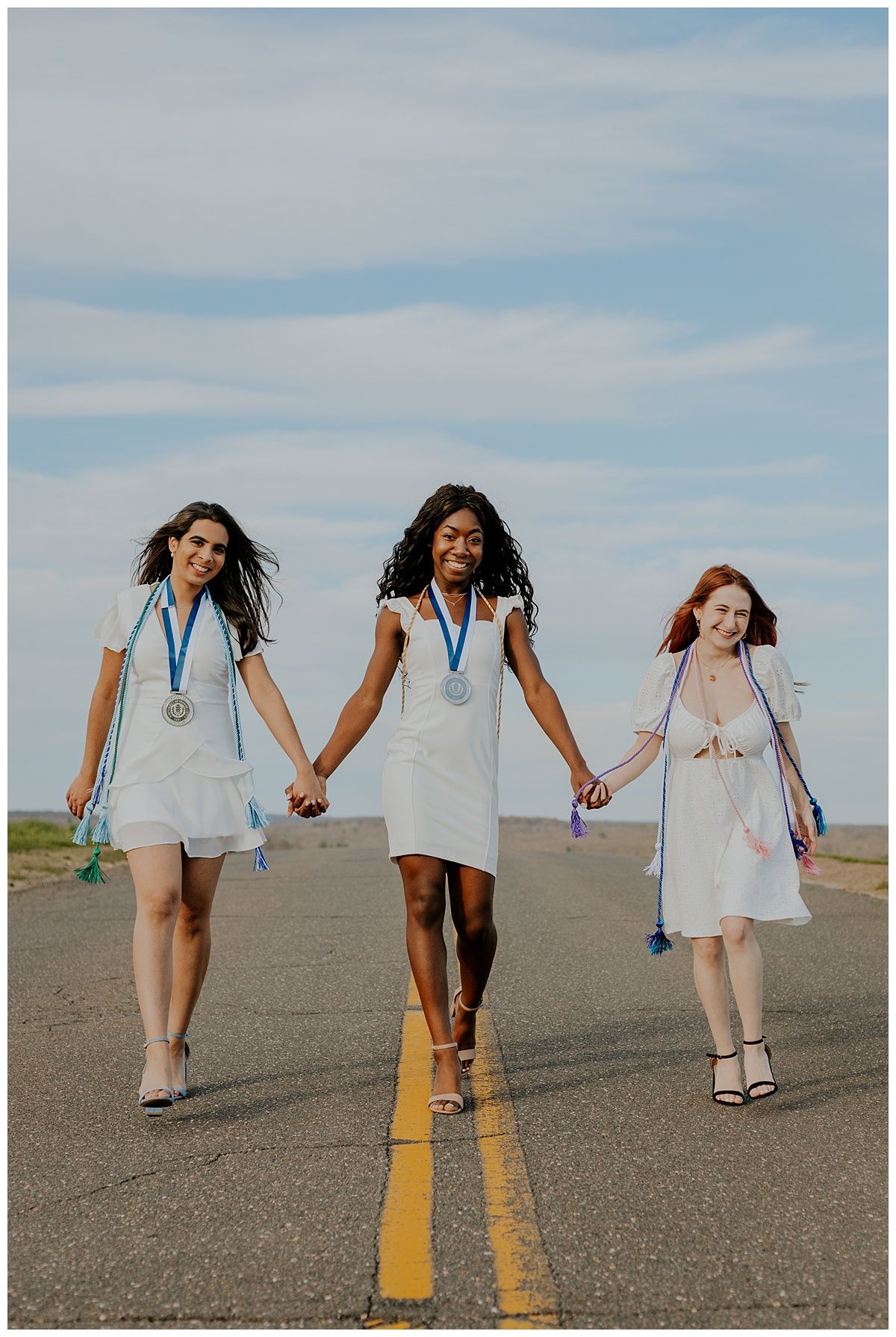 Three graduates holding hands walk down center of empty road wearing medals and white dresses.