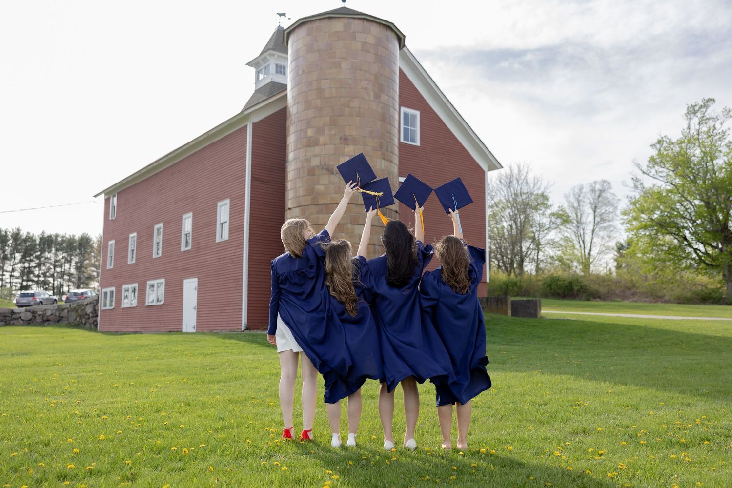 A group of graduates in blue caps and gowns jumping together in front of a red barn on a grassy lawn.