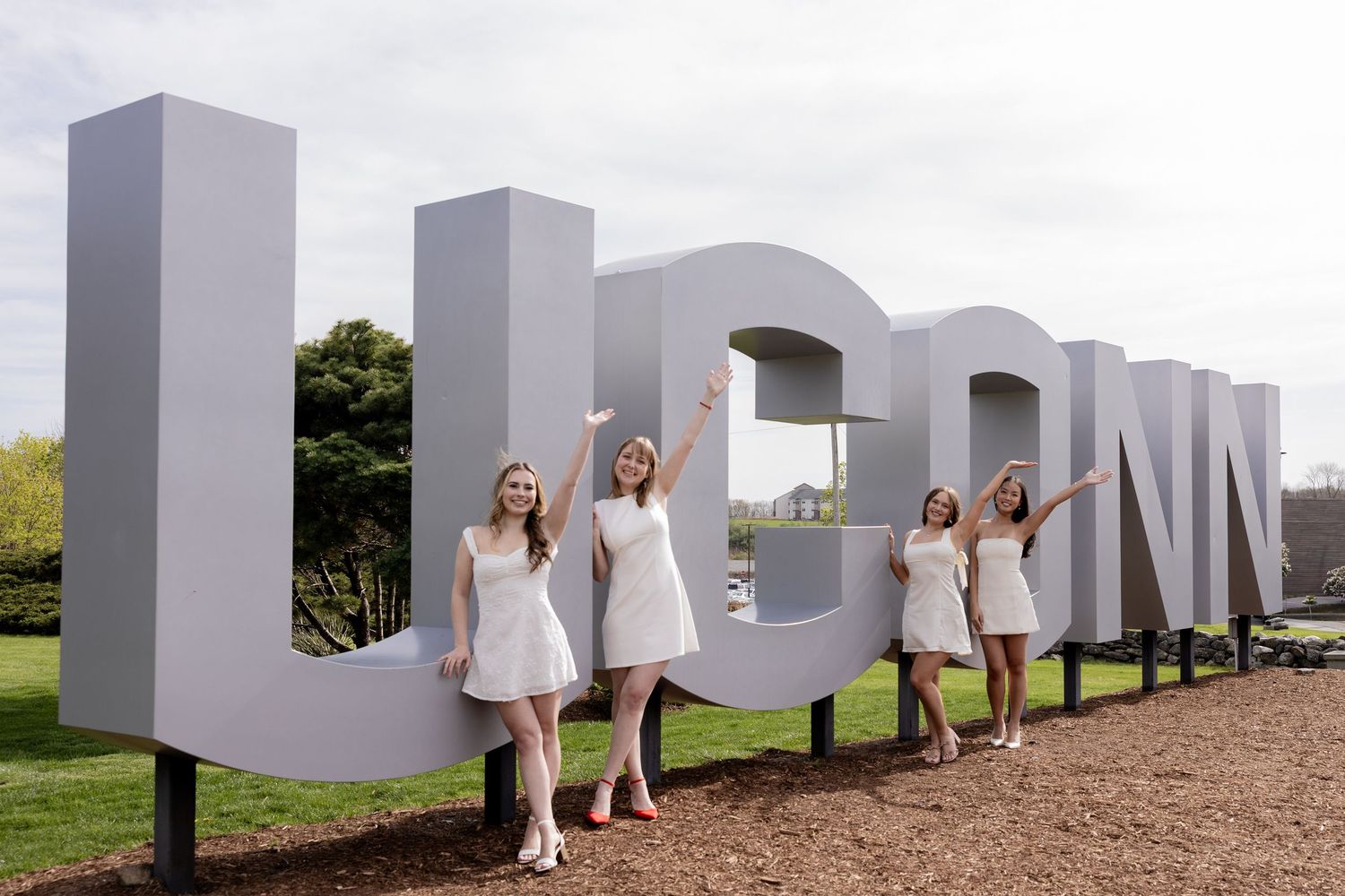 People in white dresses pose playfully along large white UCONN sign letters outdoors during a sunny day.