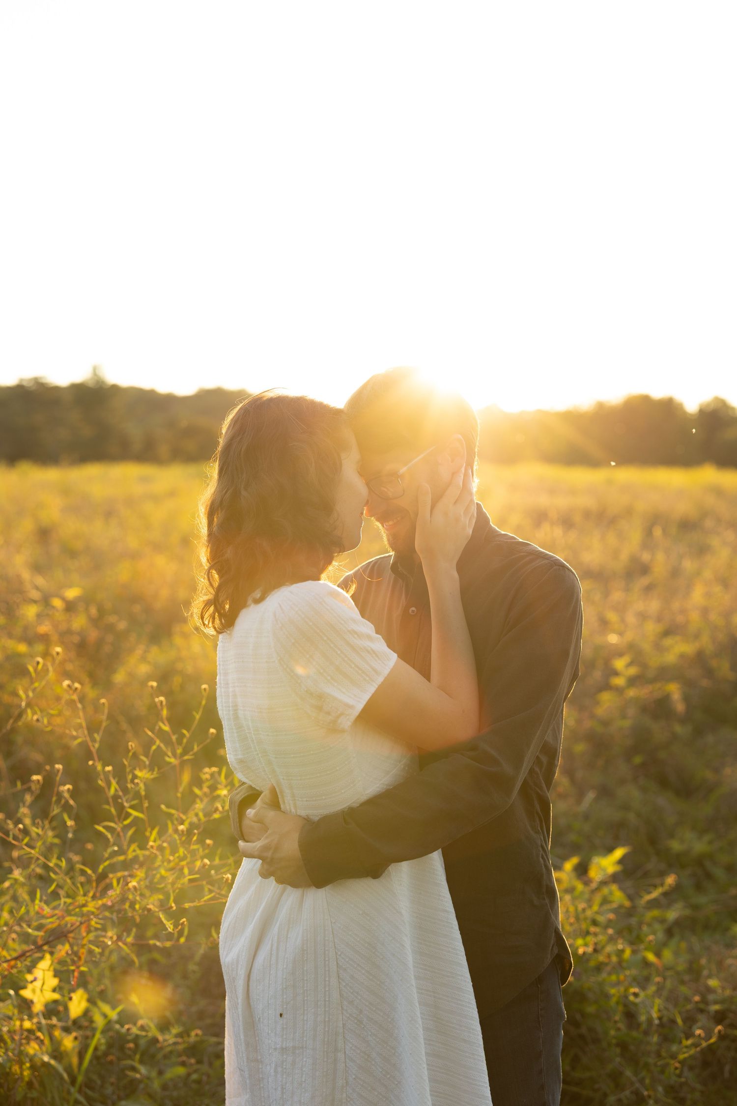A couple embraces in a sunlit meadow during golden hour as sunlight creates a dreamy backlit glow through wildflowers.