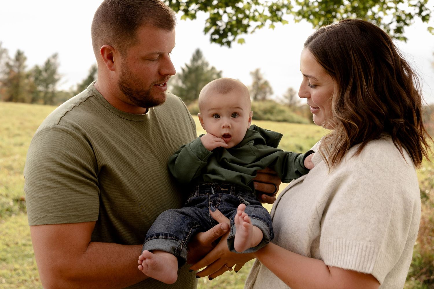 A couple holds their baby while sitting in a grassy field during a golden hour photoshoot.