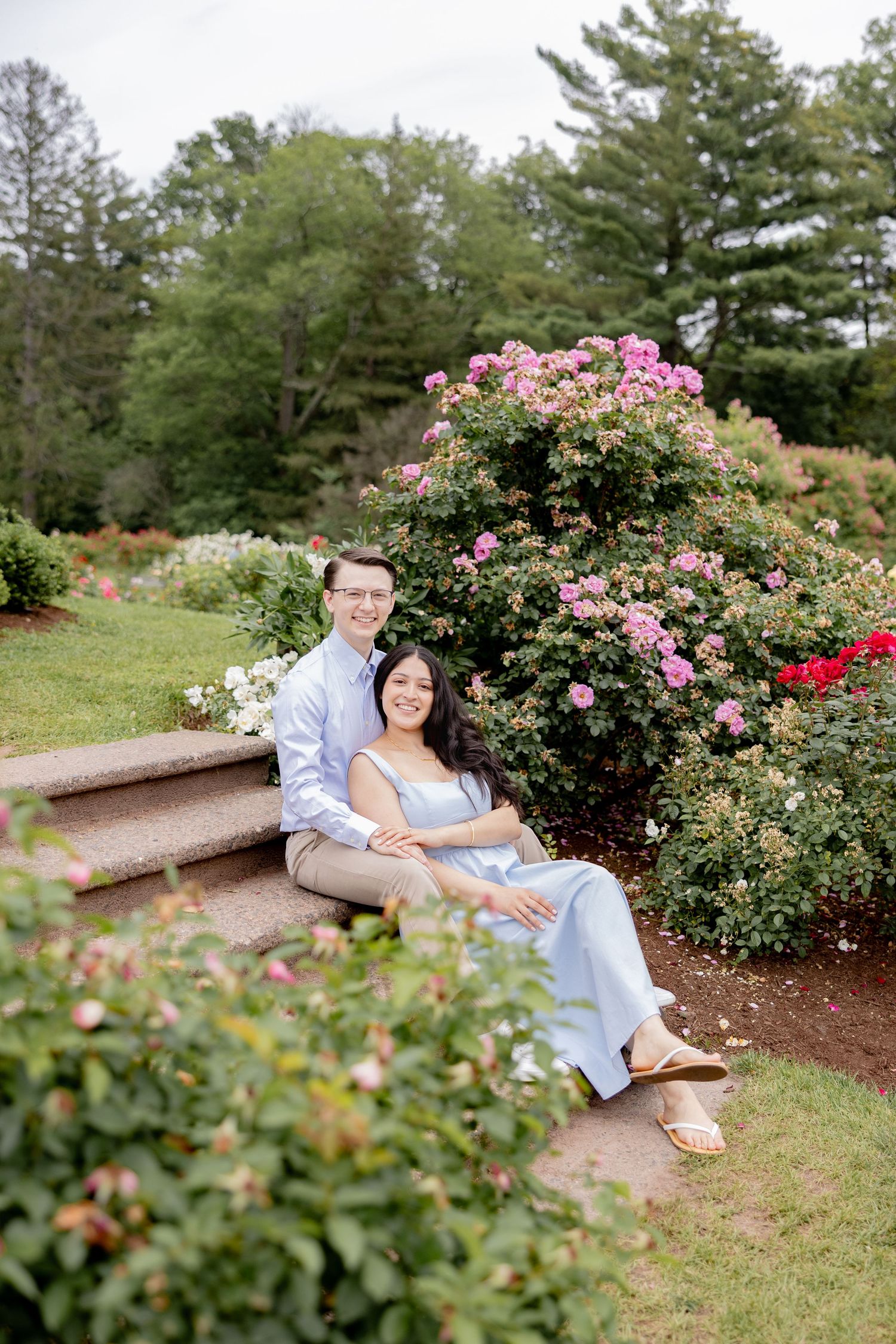 A couple sits together on a stone bench surrounded by blooming rose bushes in a peaceful garden setting.