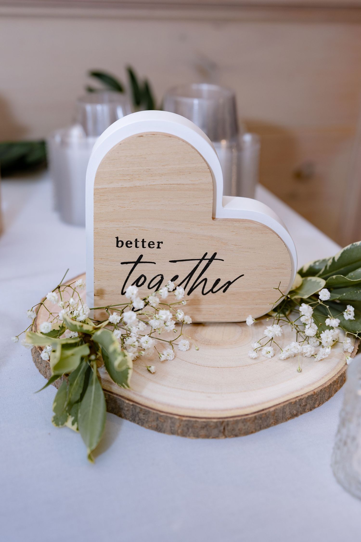 Wooden heart-shaped wedding decor with 'better together' text adorned with delicate white flowers on a rustic wood slice.