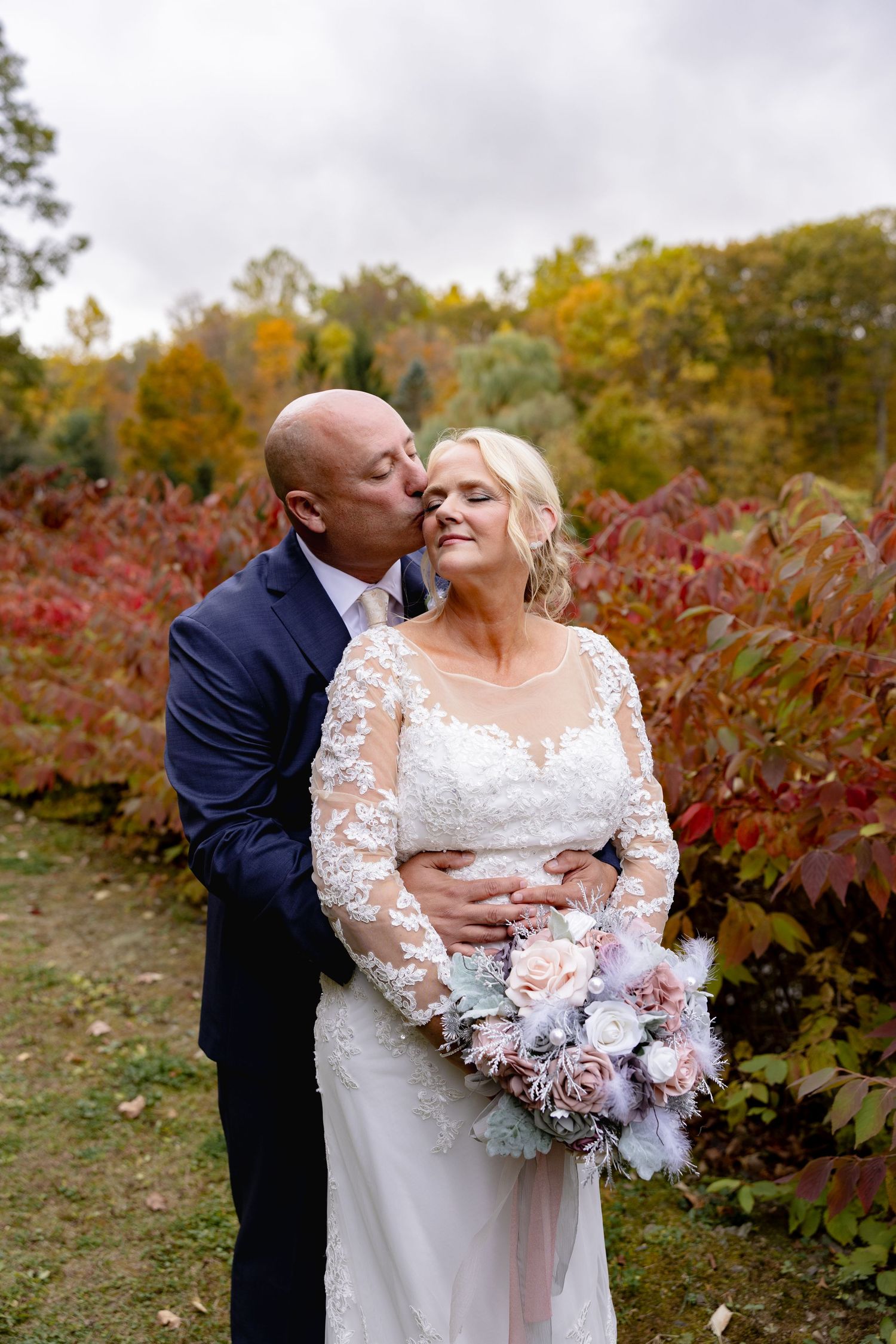 A couple shares a romantic moment in front of autumn foliage with red leaves and fall colors in the background.
