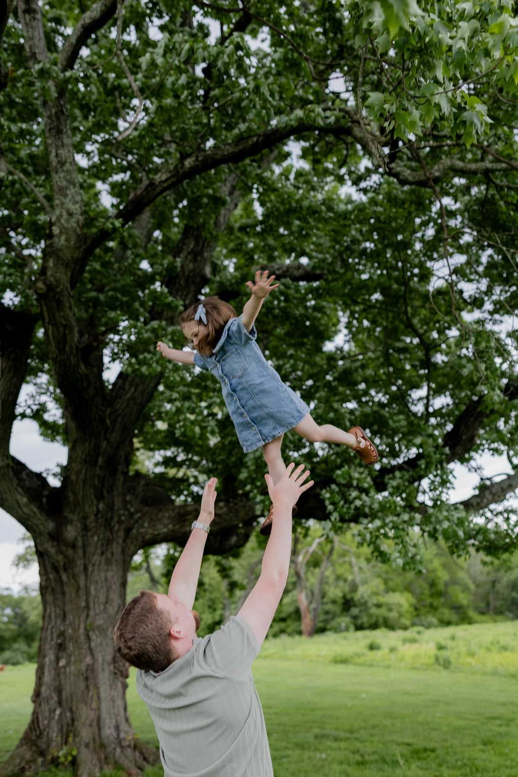 Playful outdoor scene of a child being tossed in the air near a large tree in a grassy field.