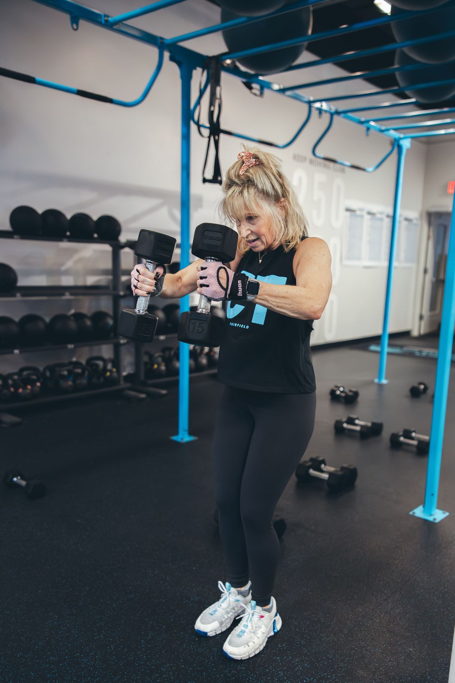 Woman in black workout attire performing dumbbell exercises in a gym with blue accents.