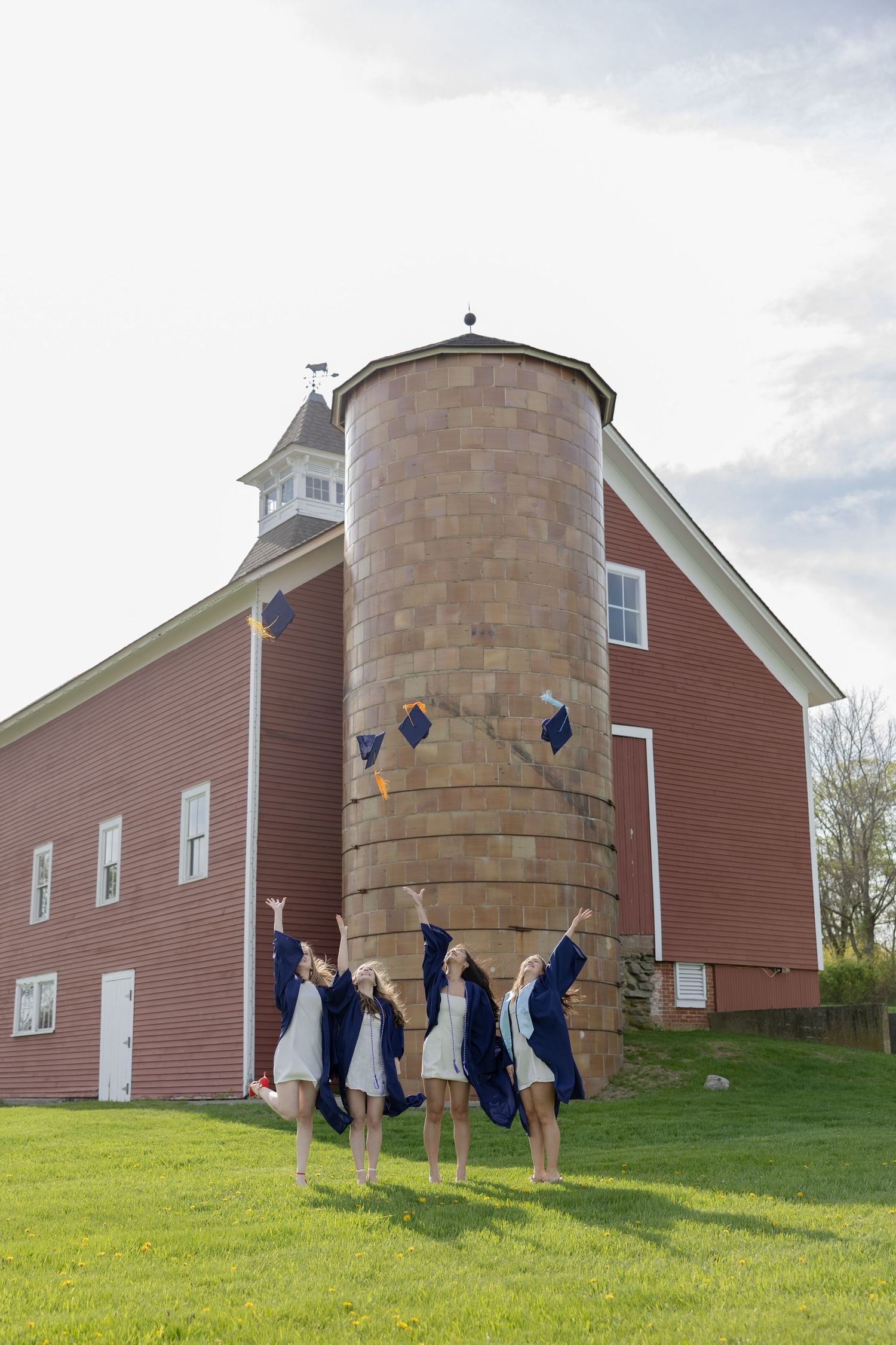 Four graduates in blue caps and gowns jump simultaneously in front of a red barn with a tall silo on a sunny day.