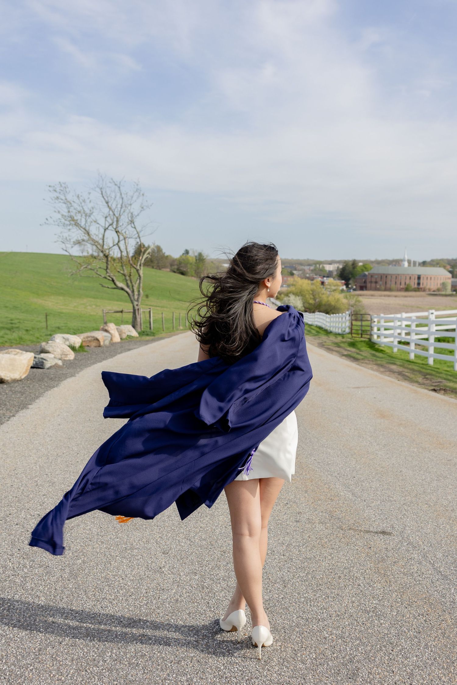 A flowing navy blue jacket billows in the wind on a country road lined with white fences and rolling green hills.