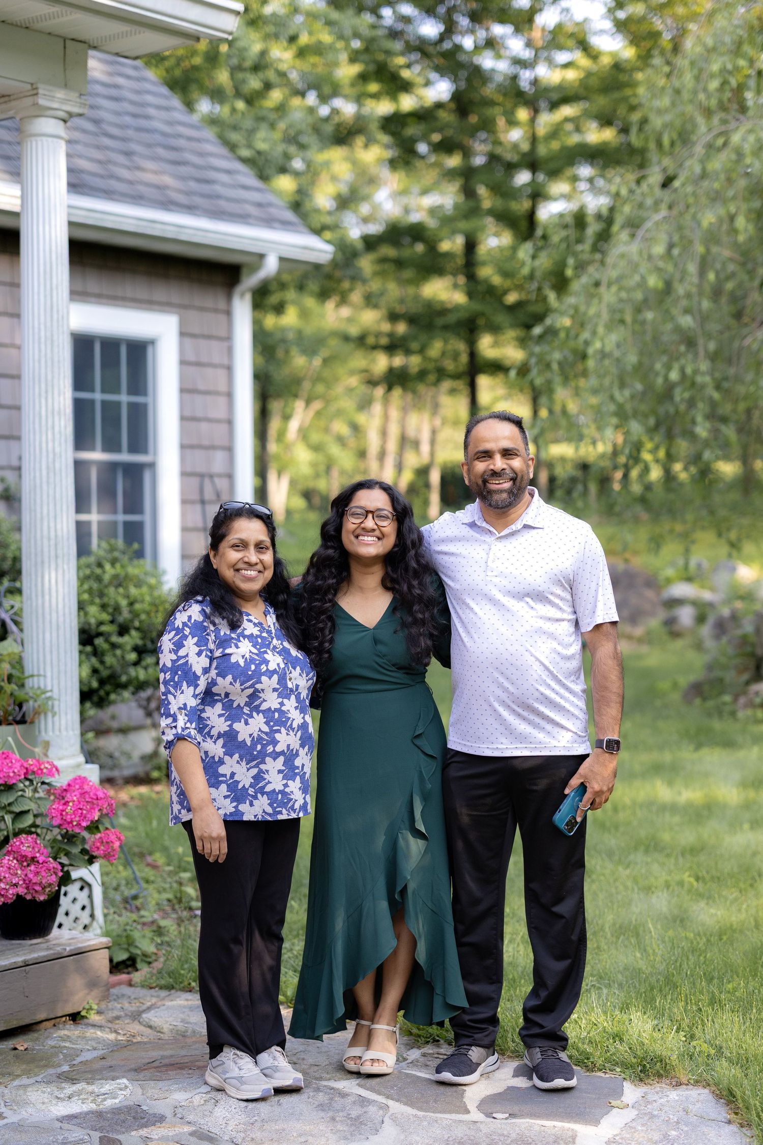 Three people stand together smiling on a stone patio outside a house with pink flowers and greenery in the background.