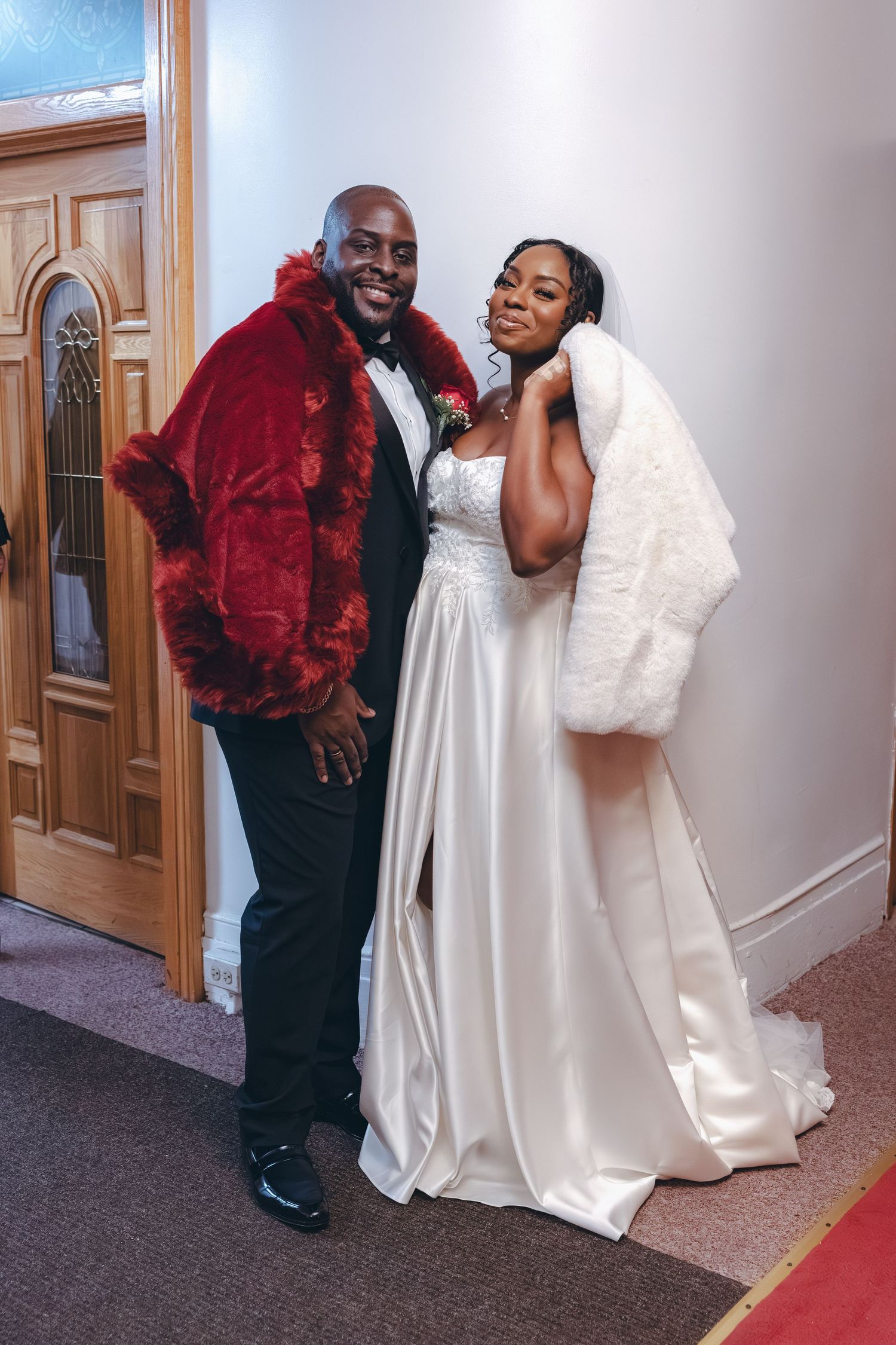 Wedding couple poses together with bride in white gown and groom wearing red velvet jacket.