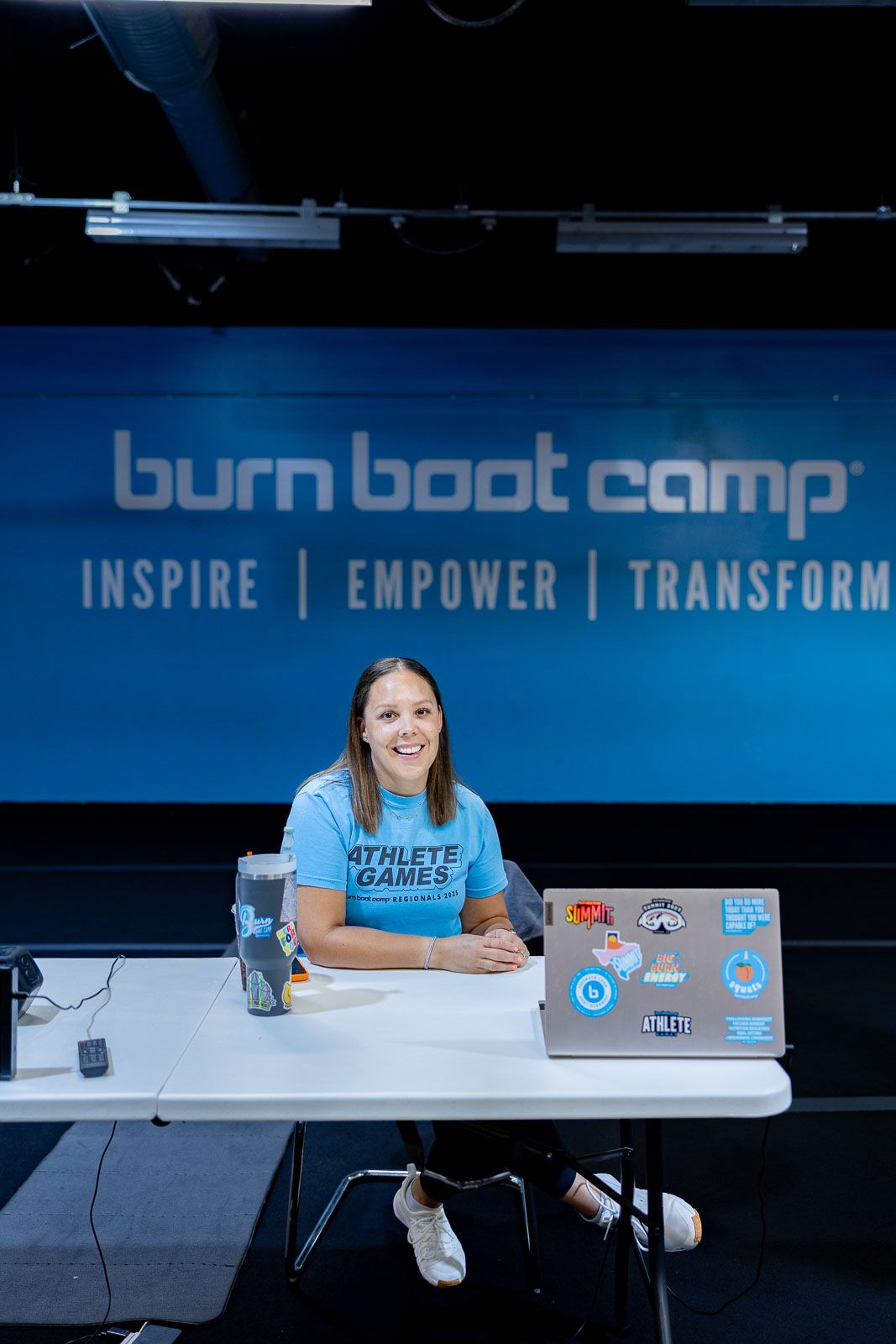 A person sits at a table with a laptop and water bottle at a Burn Boot Camp event with a blue backdrop.