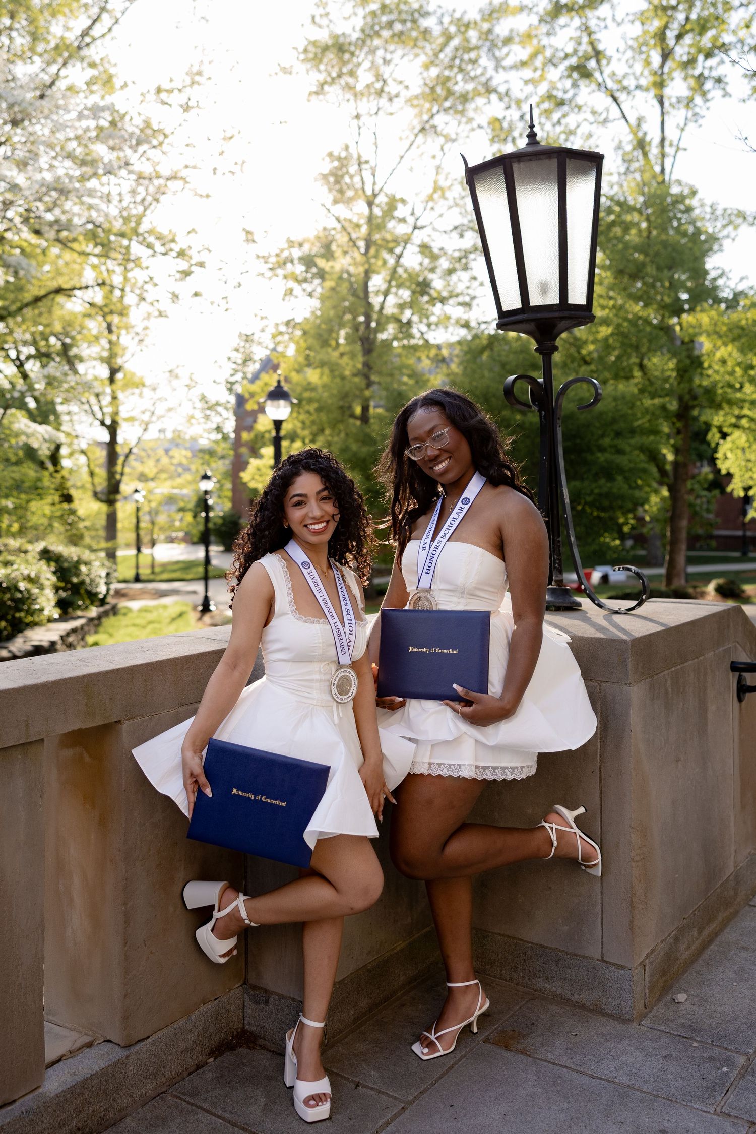 Two graduates in white dresses celebrate with diplomas while sitting on a stone wall during a sunny day.