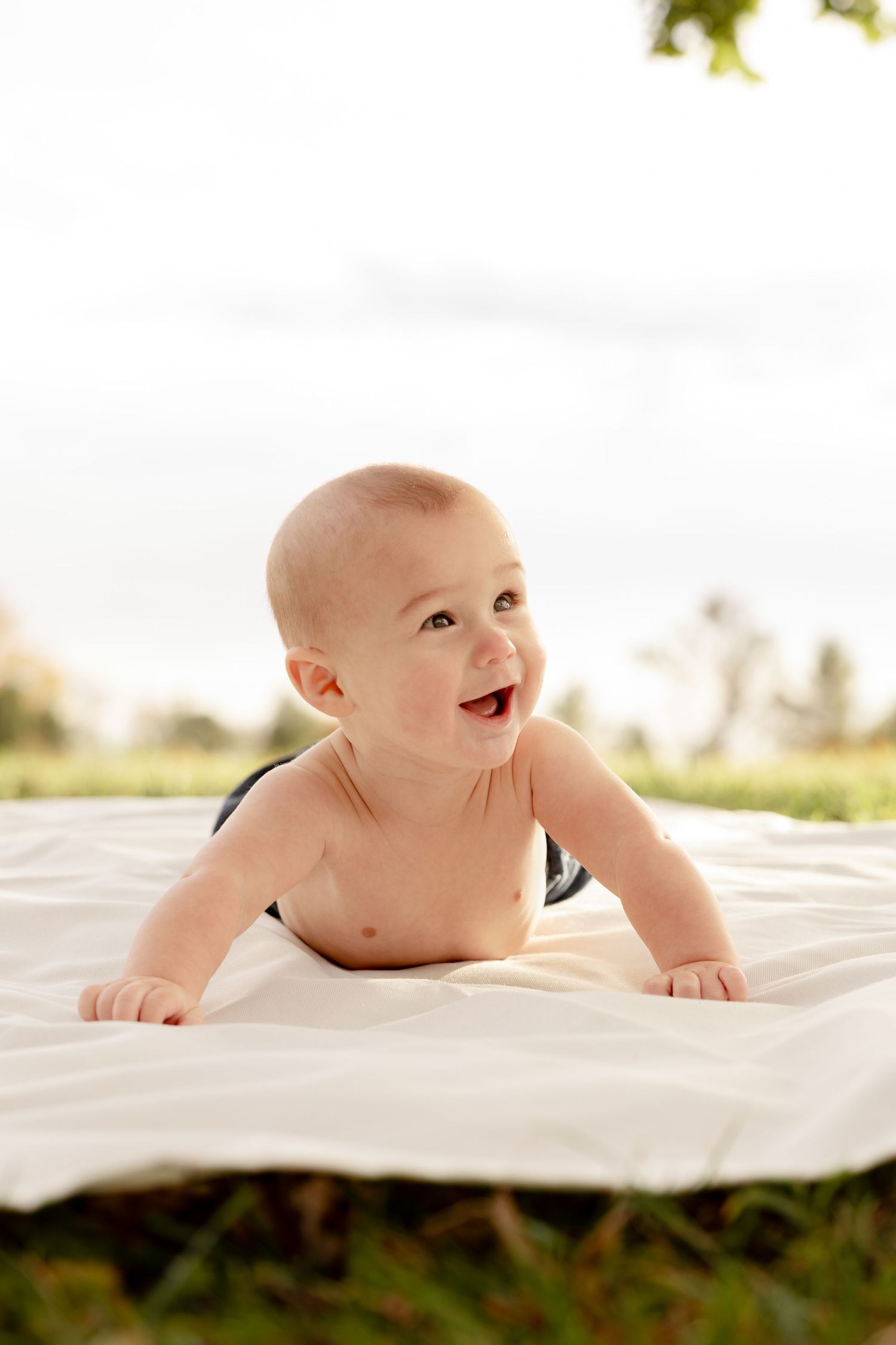 Happy baby lying on white blanket outdoors during golden hour photoshoot in nature.