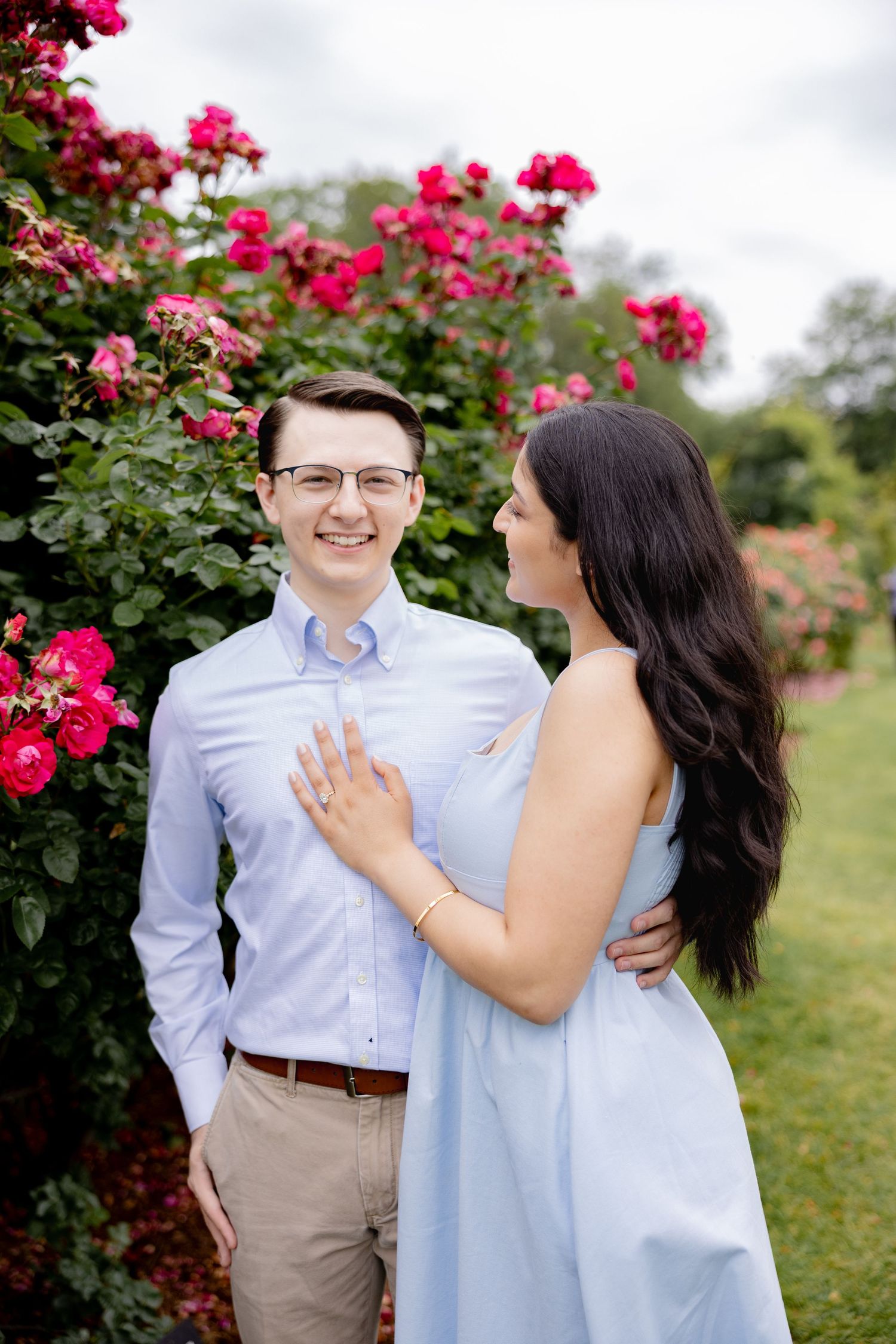 A couple poses together in front of vibrant pink rose bushes in a garden setting wearing coordinating light blue outfits.