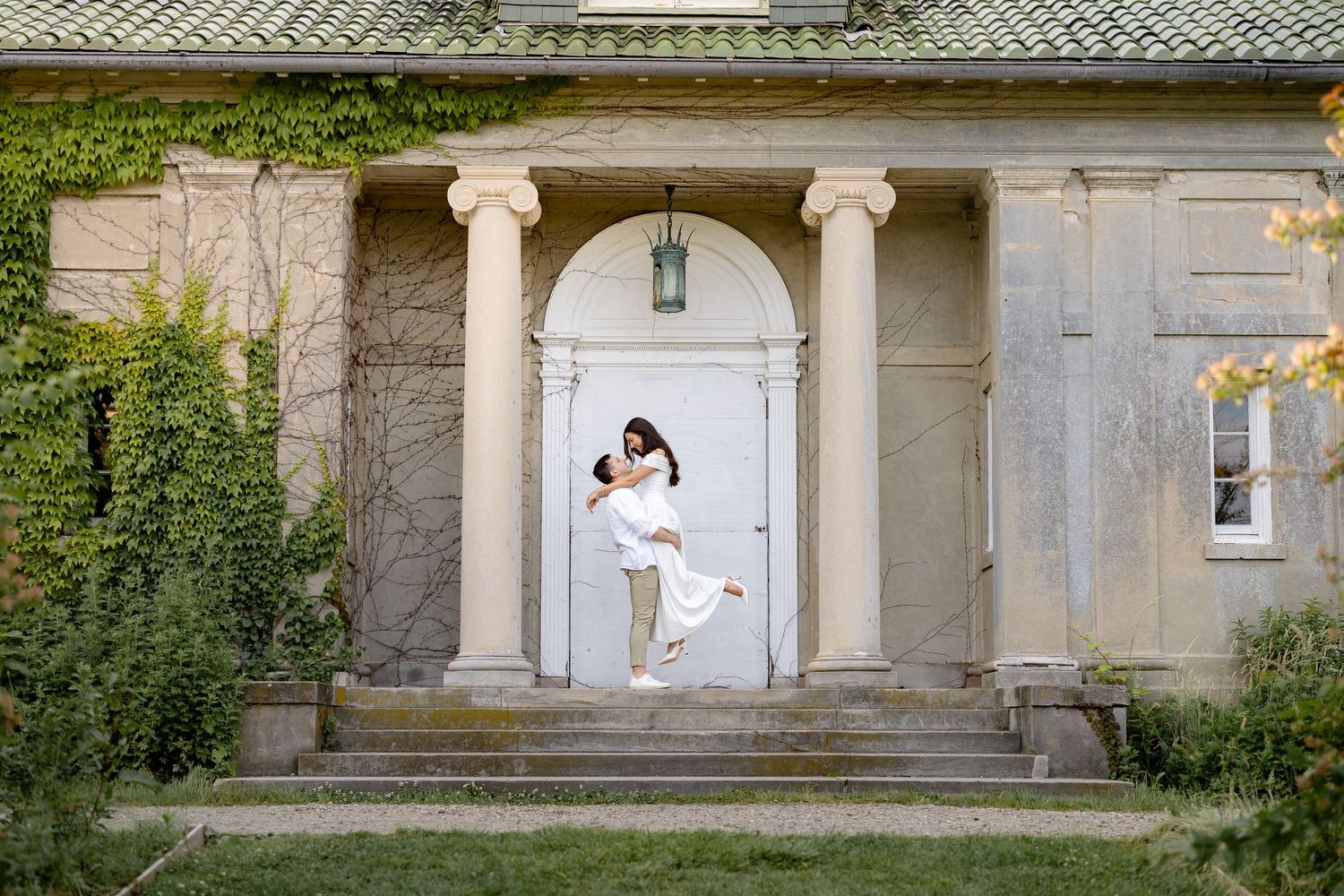 A figure in white stands elegantly in a classical stone portico entrance with columns and ivy-covered walls.
