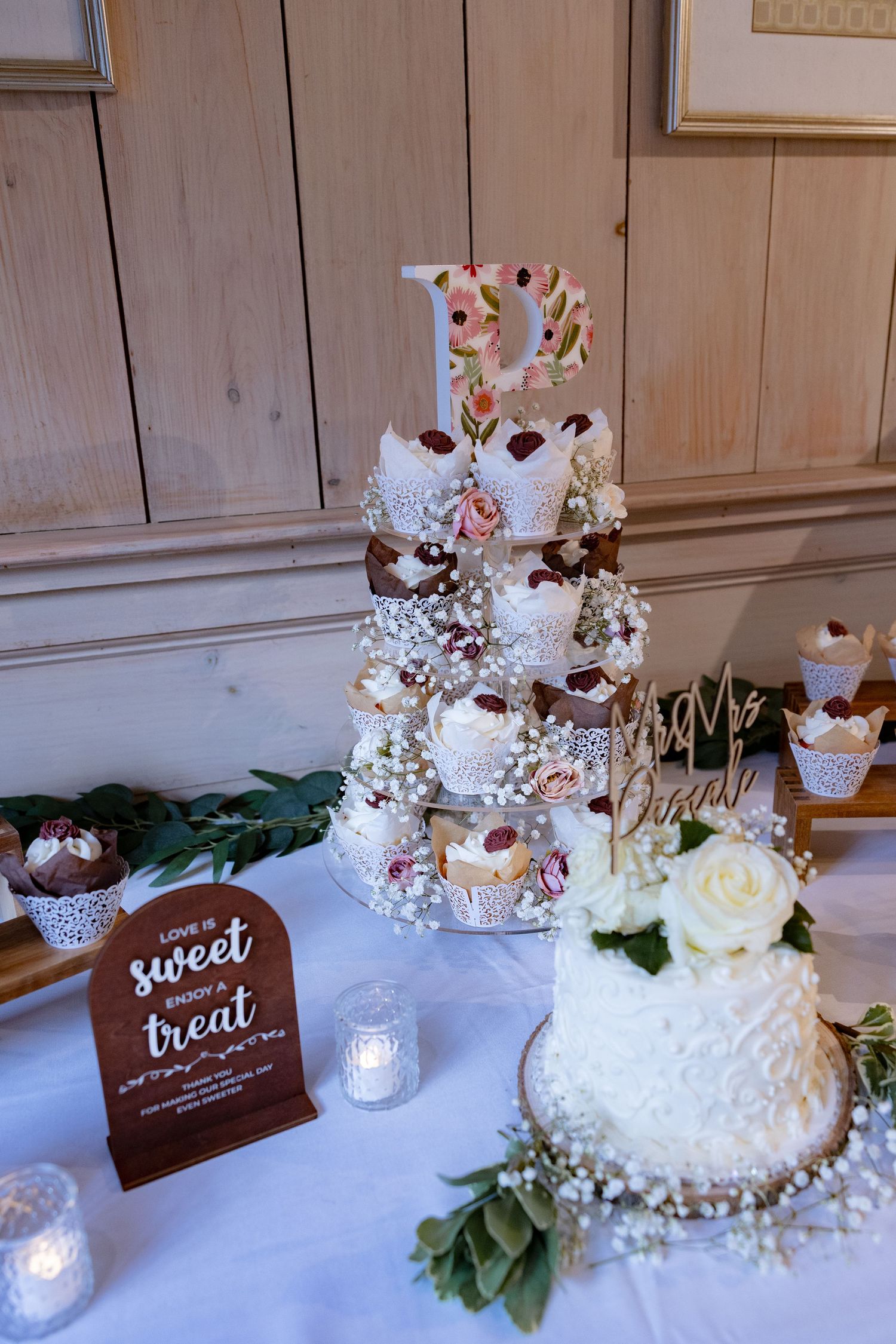 An elegant dessert display featuring white cakes and treats with wooden signage on a white tablecloth.