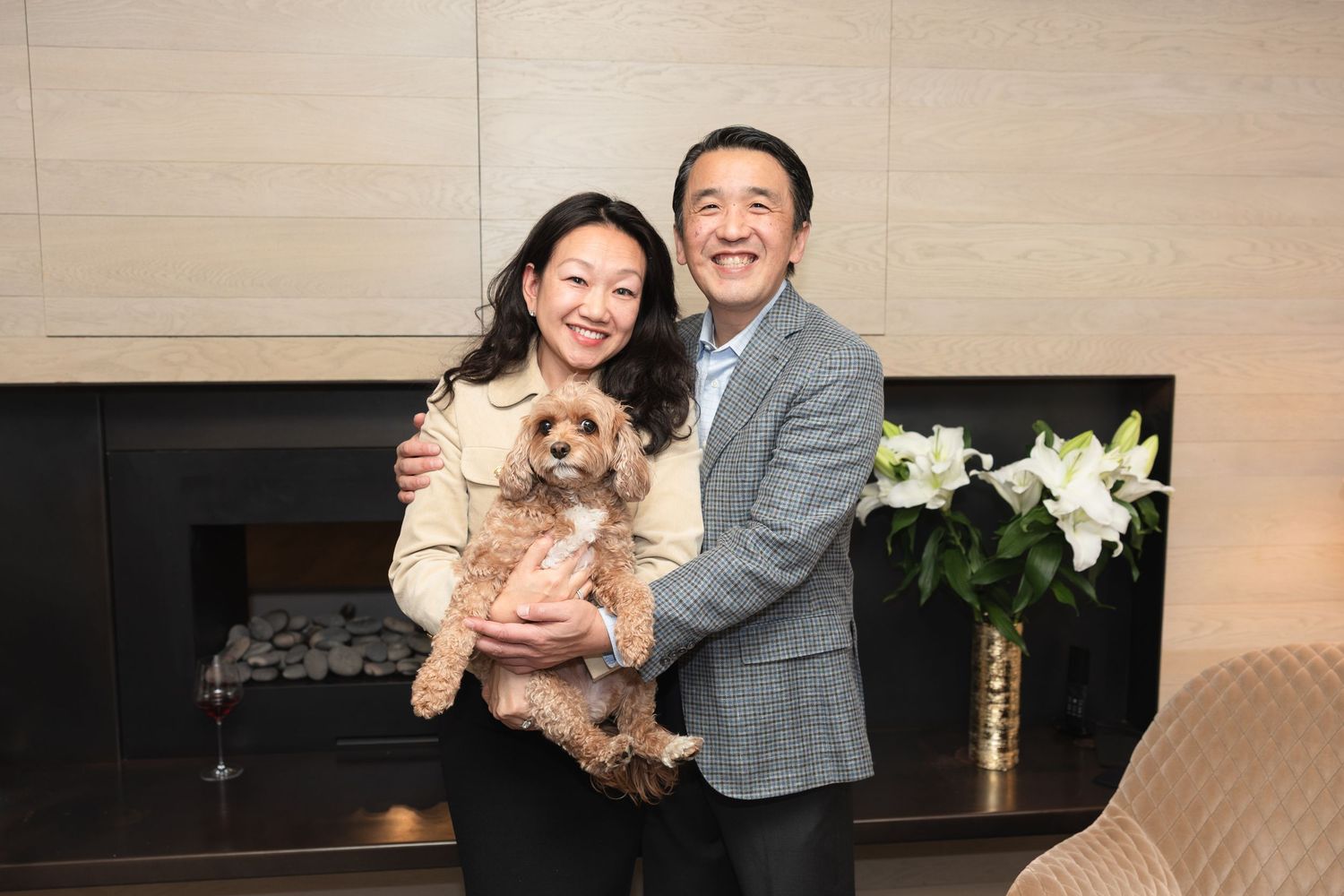 A couple poses together in a living room while holding their small fluffy dog and smiling warmly at the camera.