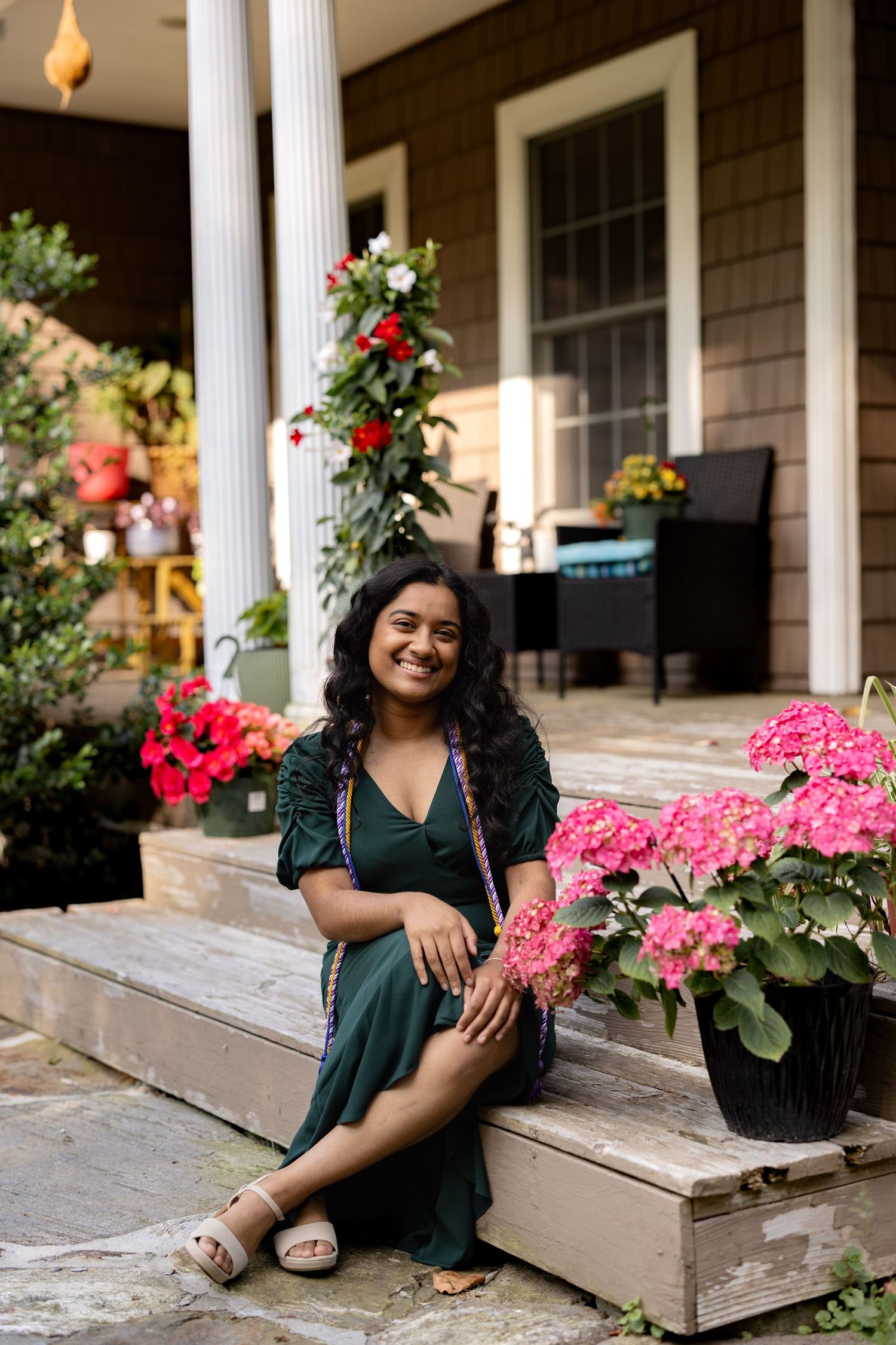 A person sits on wooden porch steps surrounded by vibrant pink flowers and climbing roses near white columns.