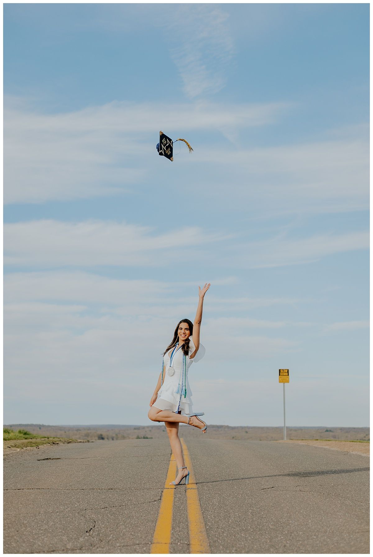 Young woman in light dress throws graduation cap on empty road with blue sky background.