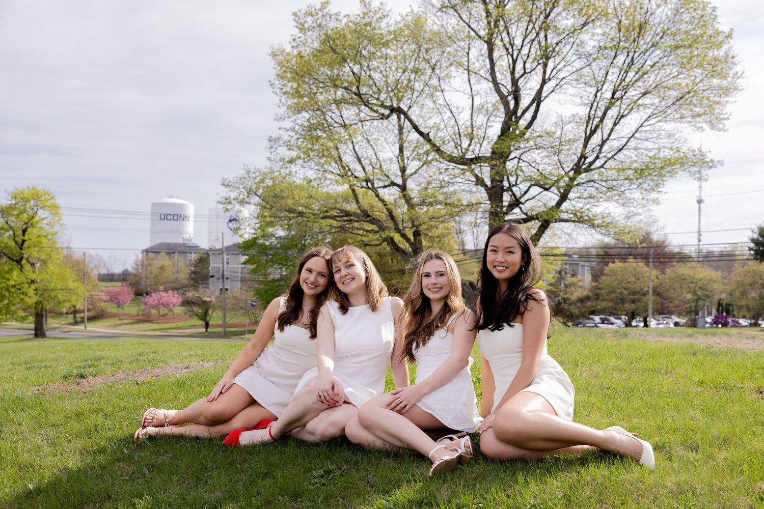 Four friends wearing matching white dresses sit together on grass with spring trees and buildings visible behind them.