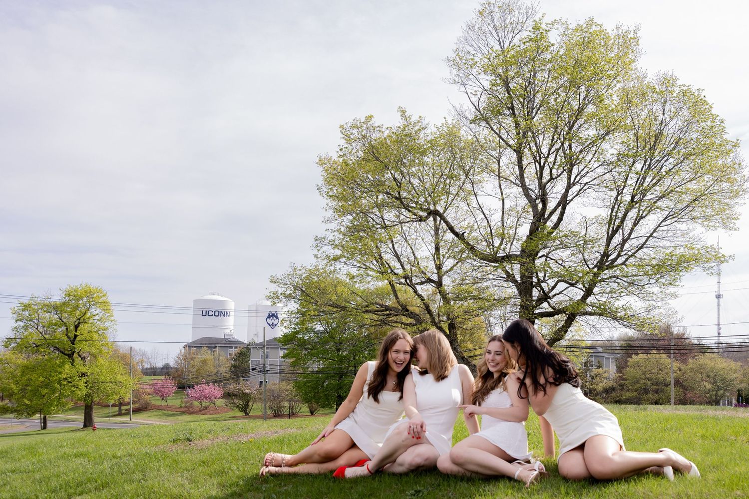 A group of friends in white dresses relax together on a grassy hill during spring with trees and buildings in background.