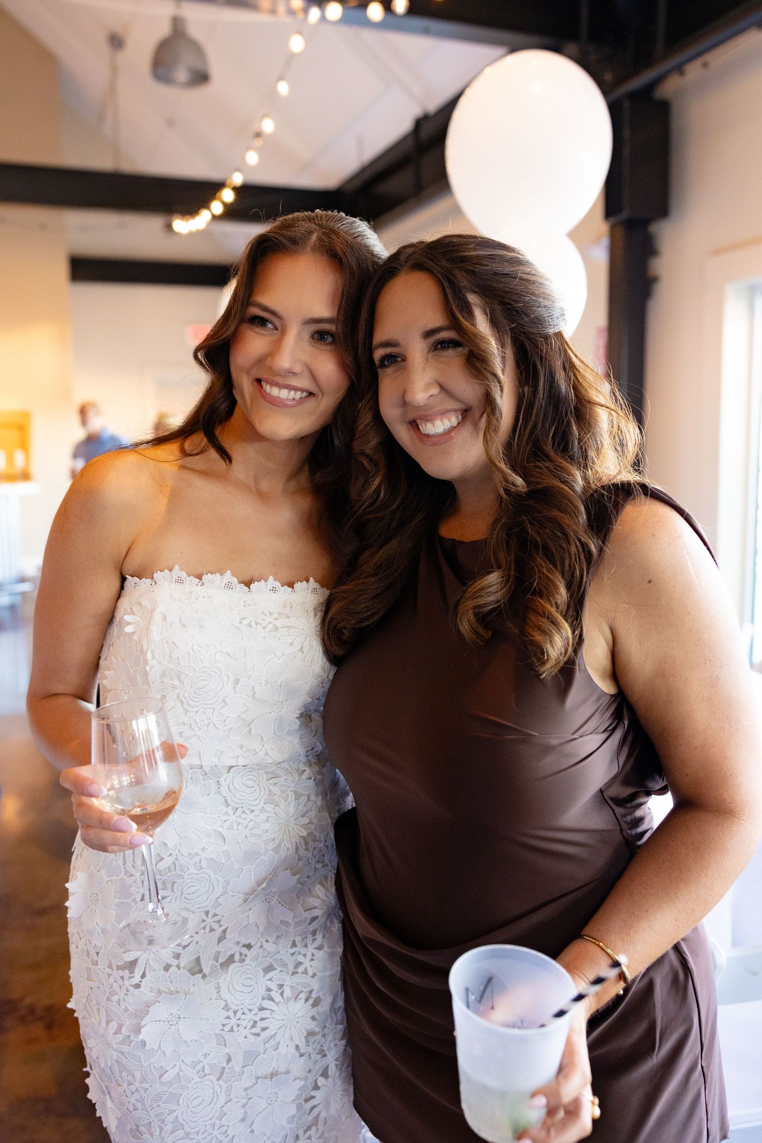 Bridesmaids wearing contrasting white lace and brown dresses pose together with drinks at a wedding celebration.