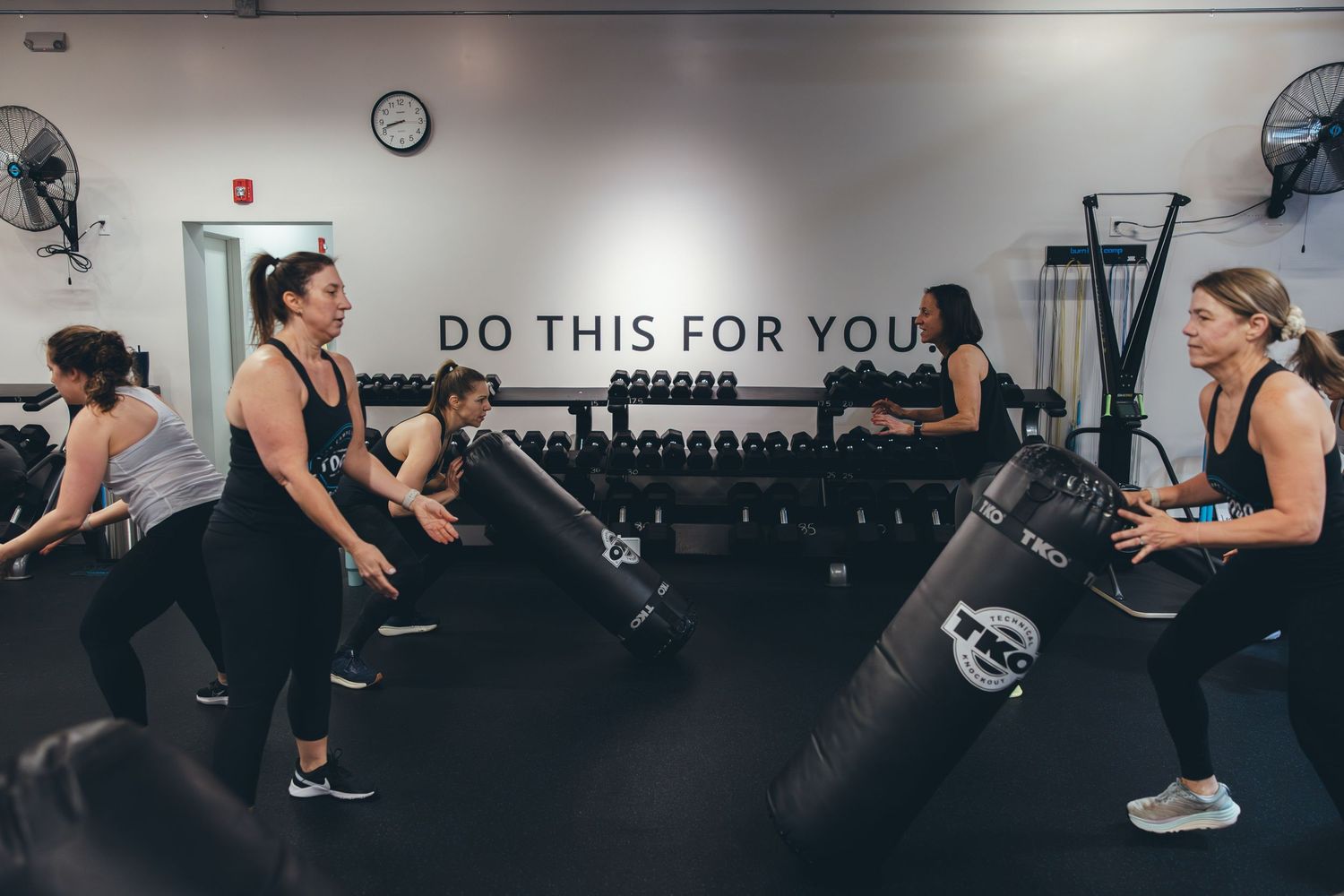 Group fitness class participants working out in a gym with 'DO THIS FOR YOU' text on wall.