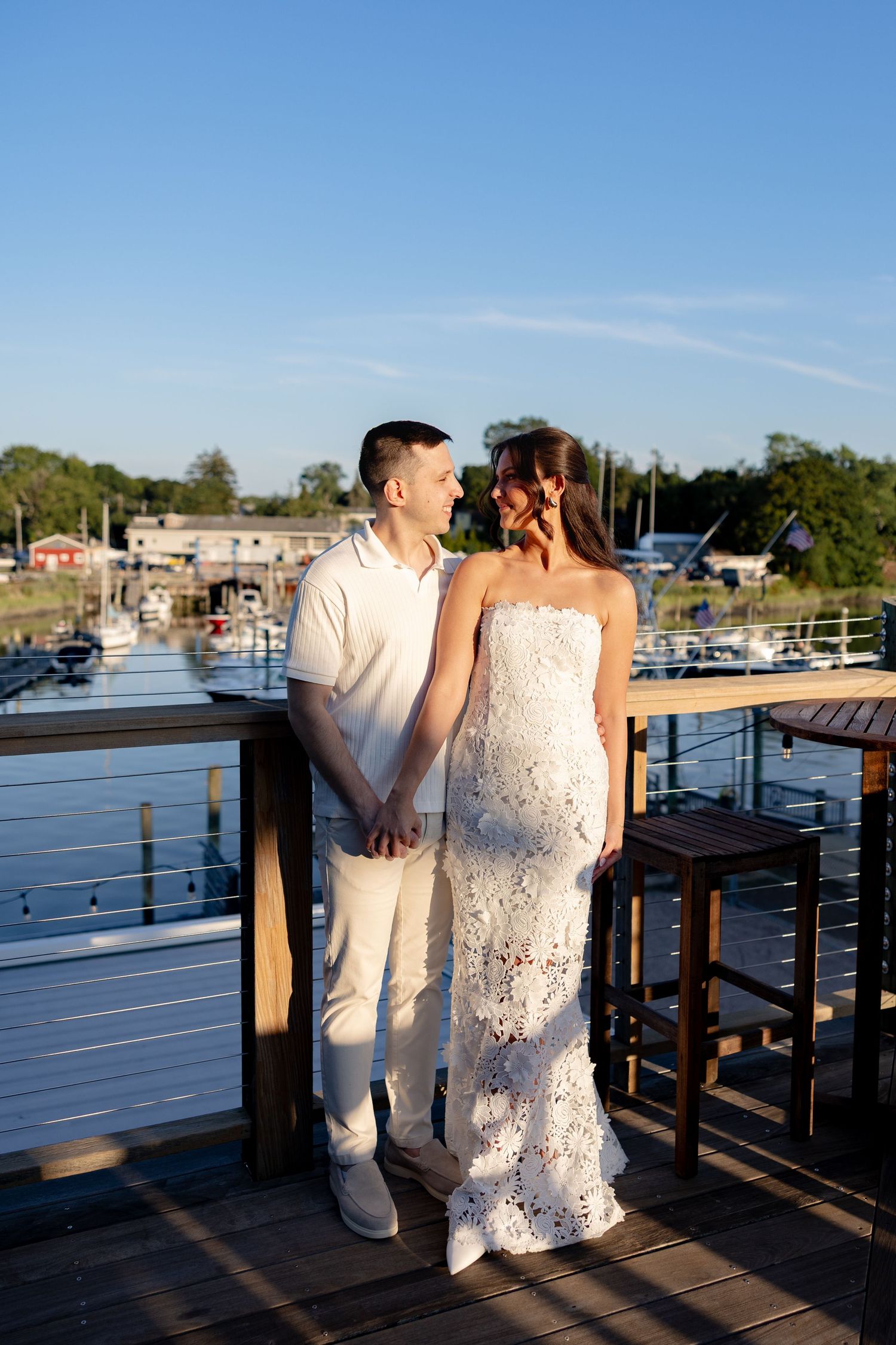 A couple in white attire shares a romantic moment on a wooden dock overlooking a marina at sunset.