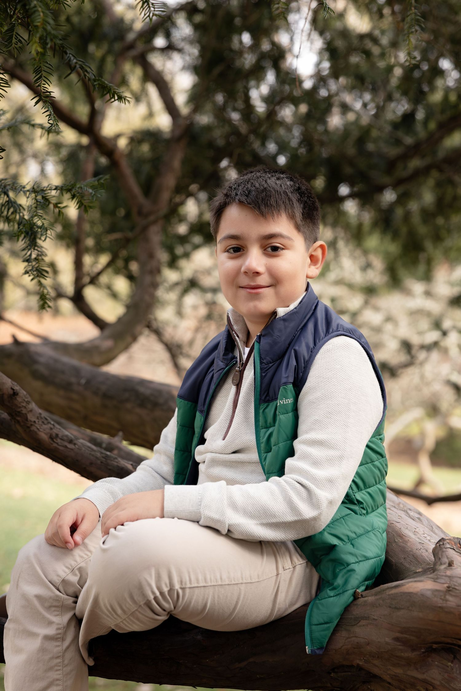 A child in a green vest and beige pants relaxes on a tree branch during an outdoor photo session.