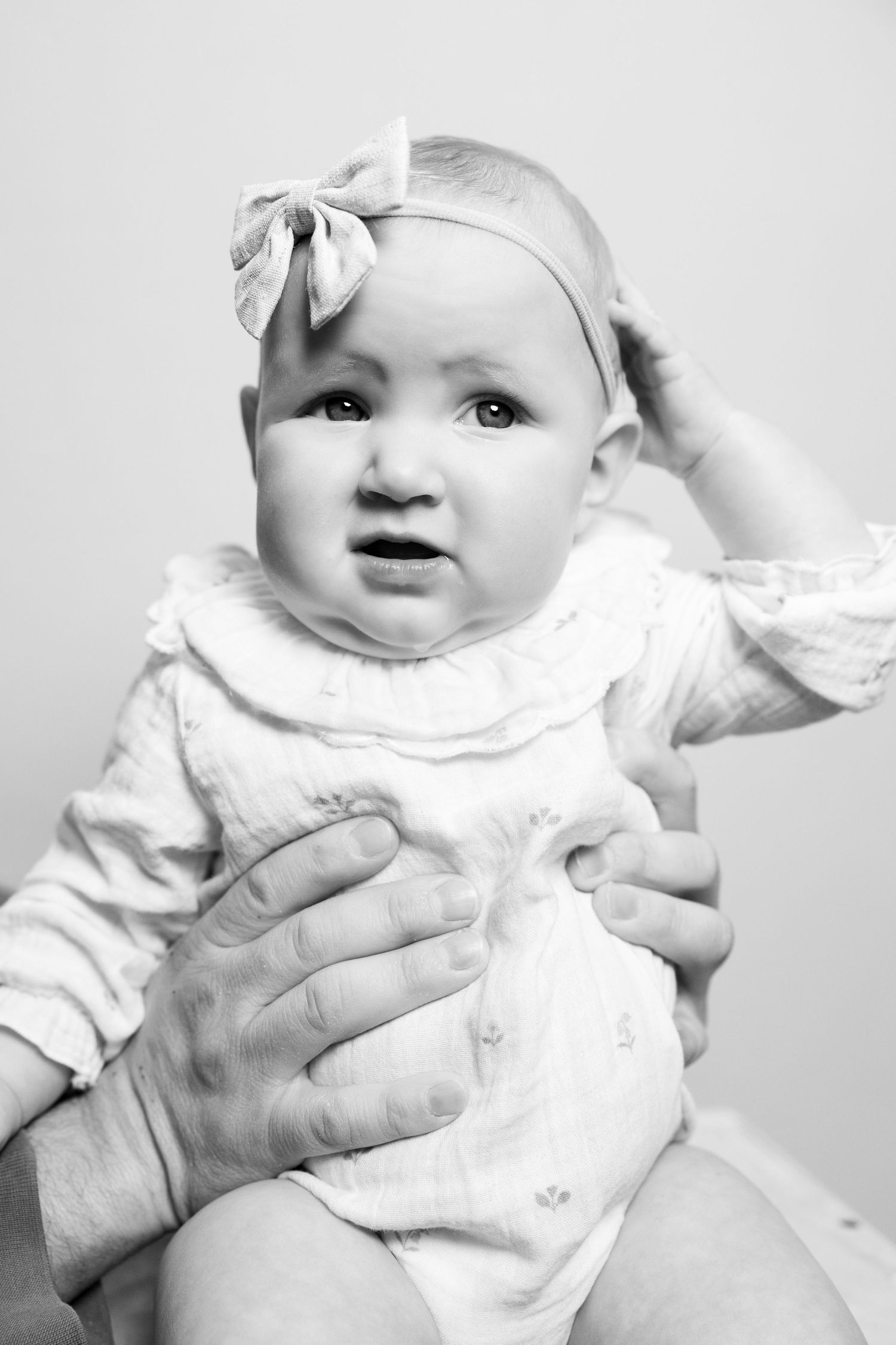 Black and white photo of an infant wearing a white outfit with bow headband being held by adult hands.