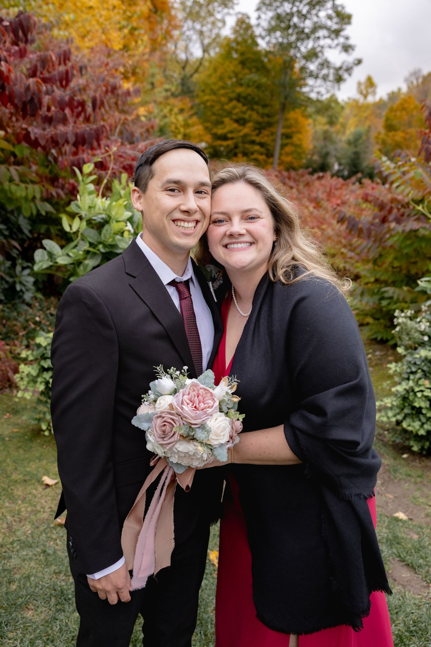 A couple in formal attire shares a joyful moment together in a garden with vibrant fall foliage in the background.